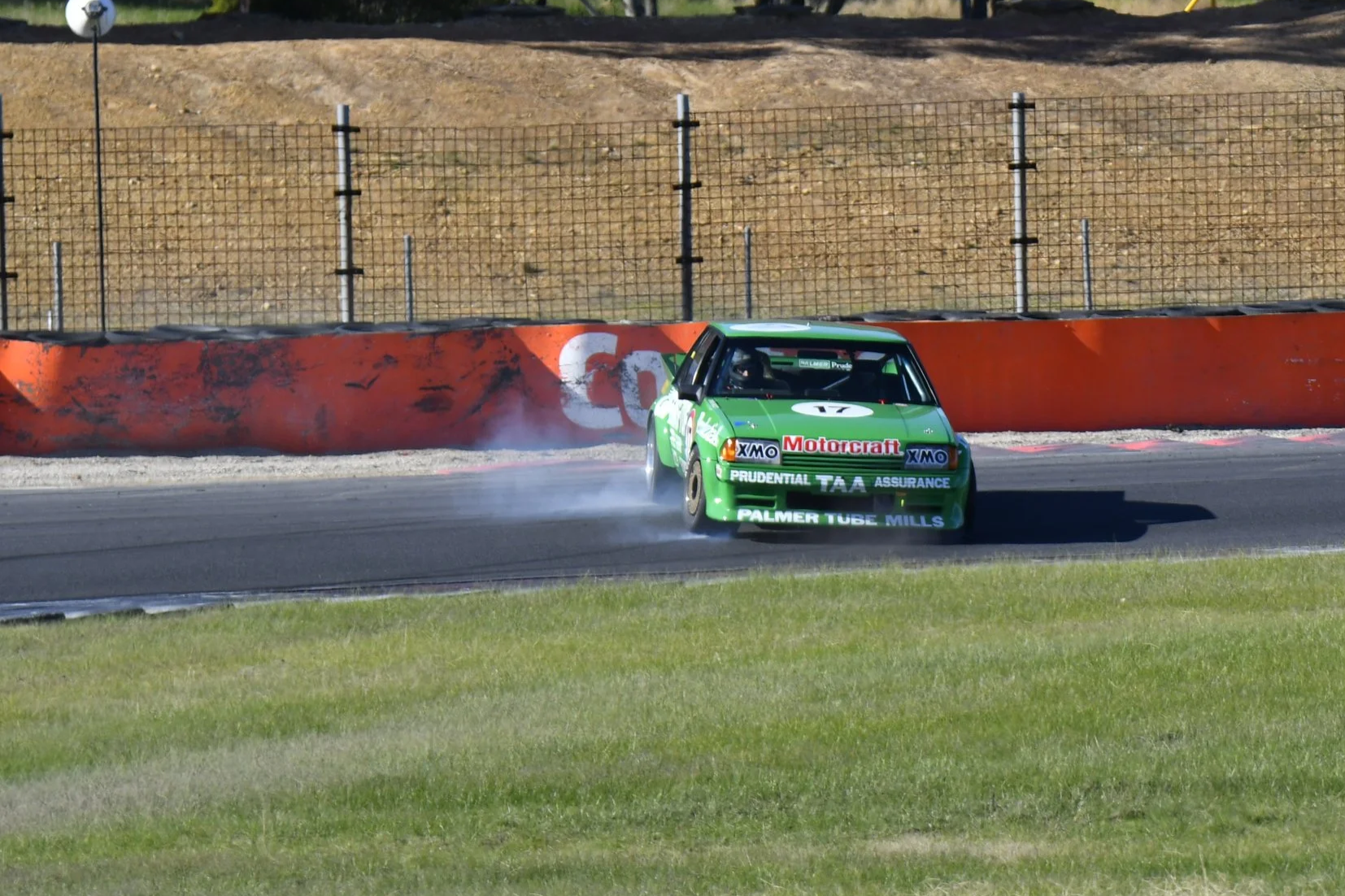 A bright green race car with sponsors' logos is drifting around a bend on a race track, creating smoke from its tires, with a red barrier and a fence in the background.