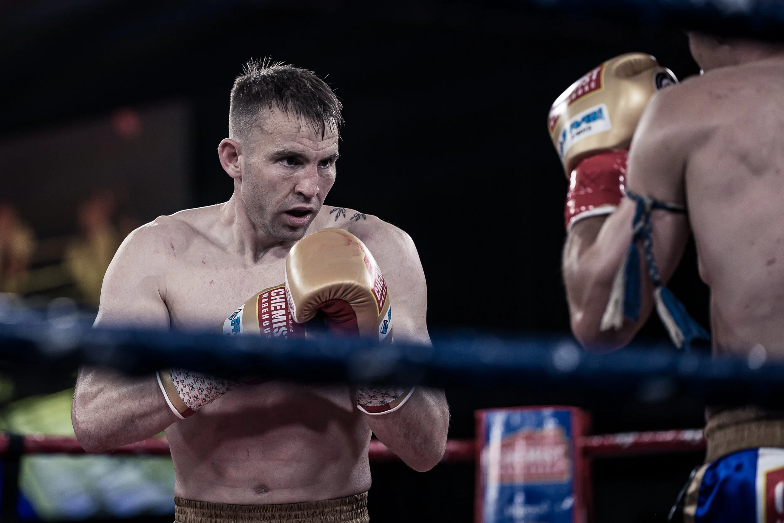 Two shirtless men with boxing gloves in a boxing ring preparing to fight, with a dark background and blurred spectators.
