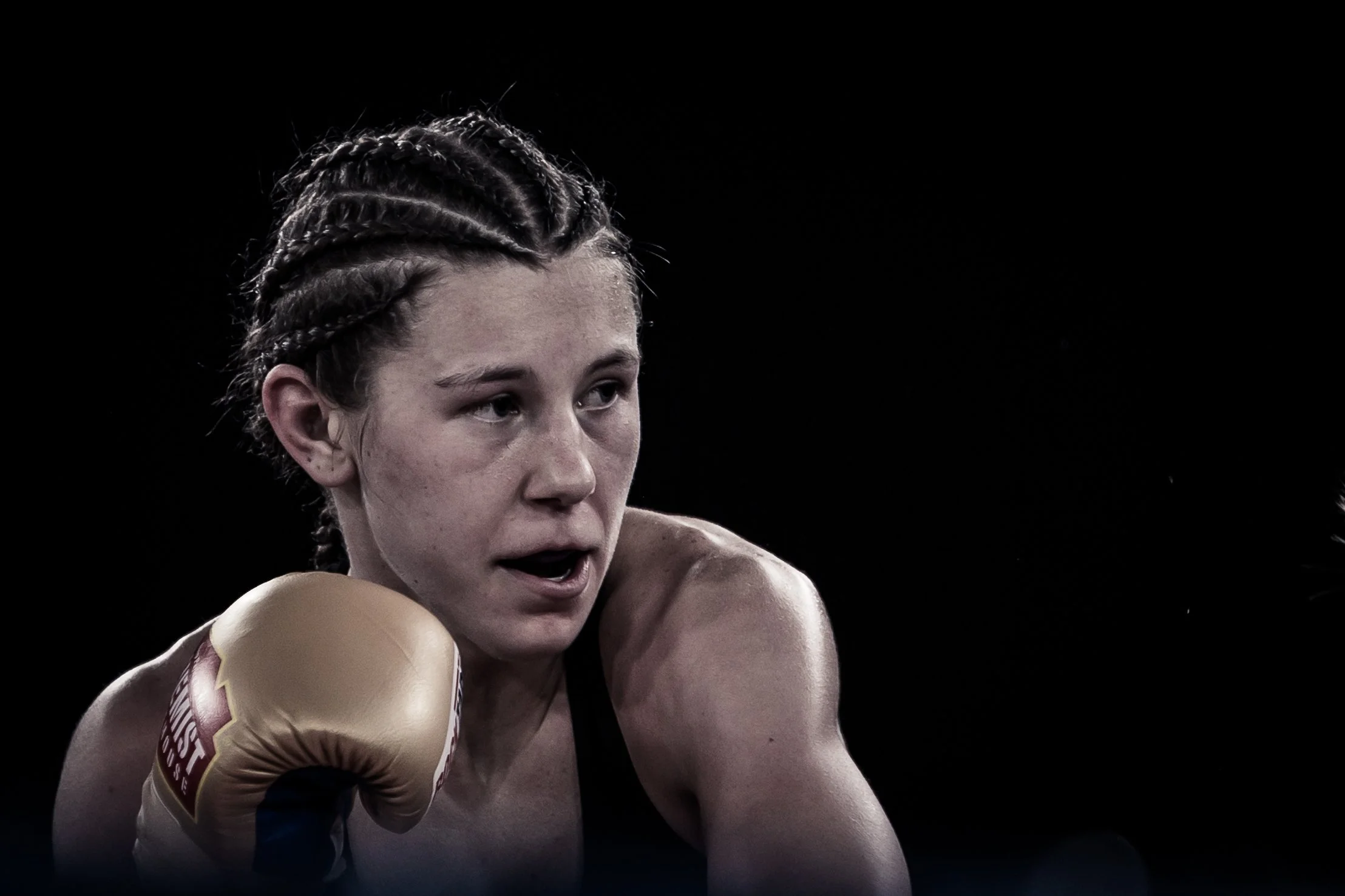 Close-up of a female boxer with braided hair in boxing gear, looking focused during a match against a dark background.