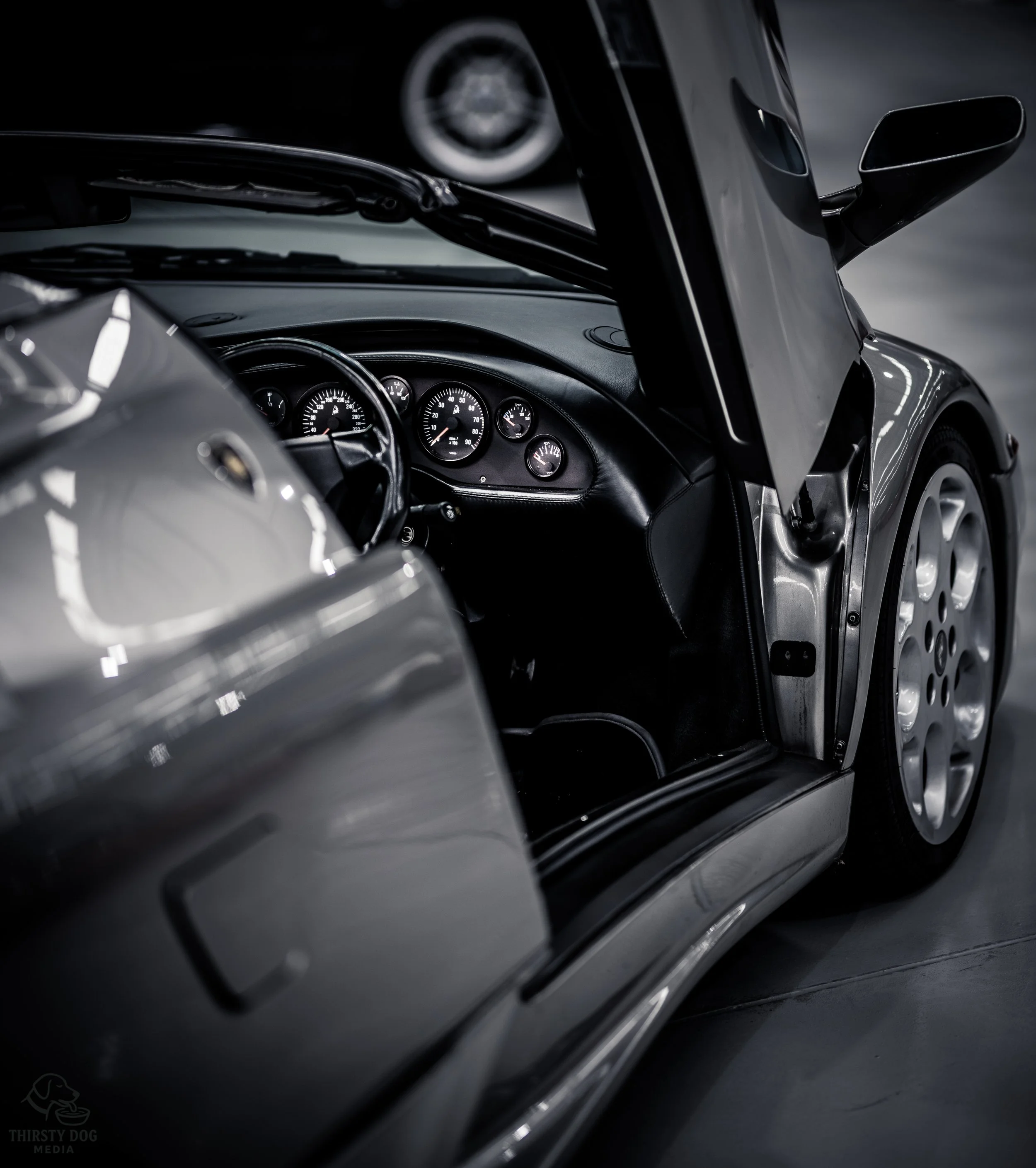Black sports car with open gullwing door, showing dashboard with gauges, set in a dimly lit garage.