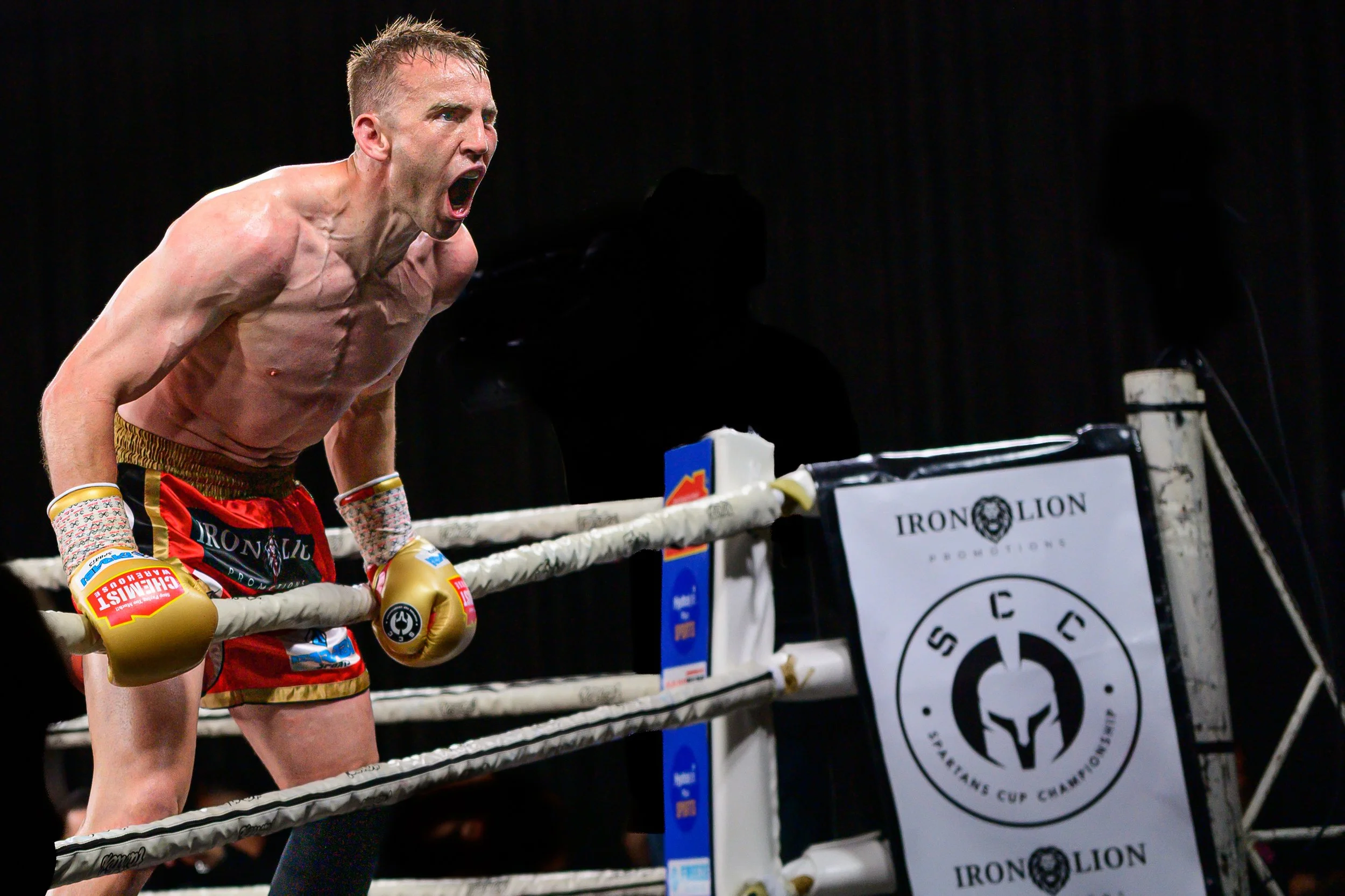 A shirtless male boxer in red shorts with gold trim, wearing gold gloves, leaning on the corner of a boxing ring, yelling with intense emotion.