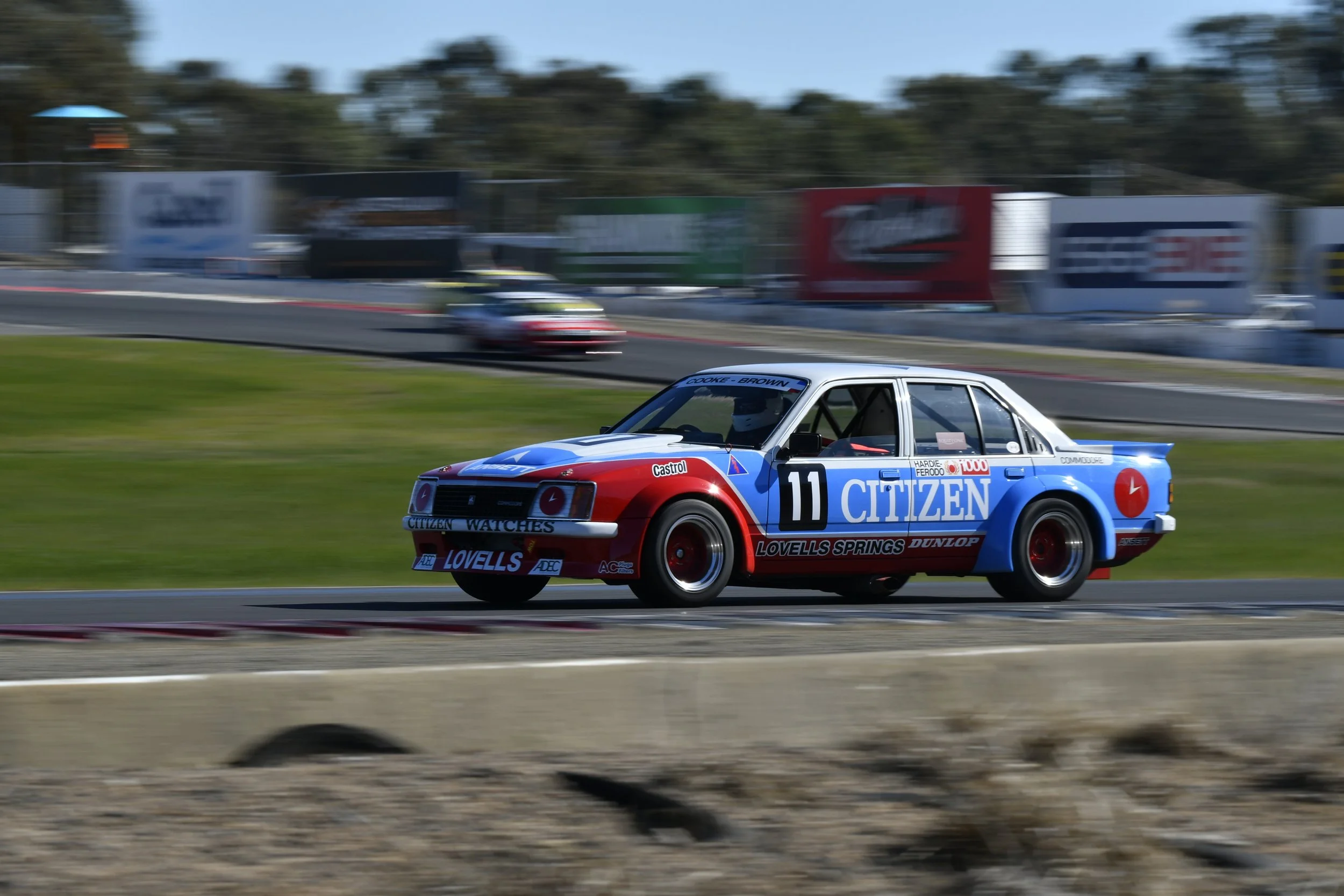 A racing car on a track, painted in blue, red, and white with number 11, with several sponsor logos including Citizen, Lovells, and Dunlop, speeding along the track during a race.