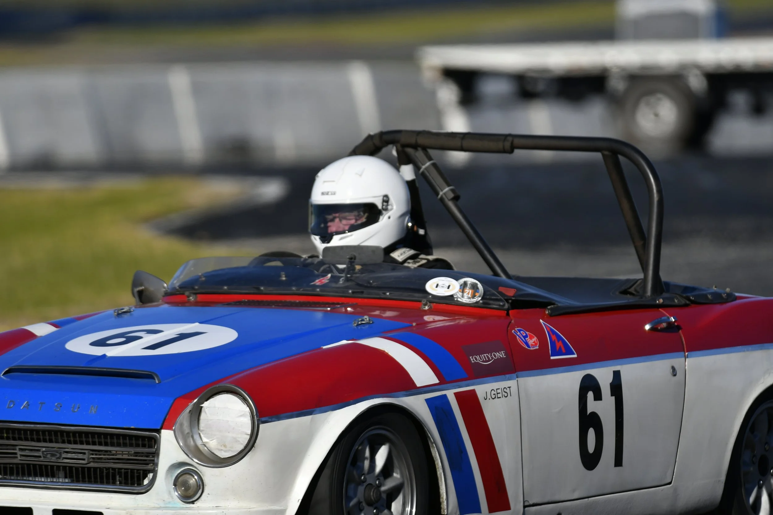 A vintage race car with a three-color red, white, and blue paint job, number 61, driven by a person wearing a white helmet, on a racing track.