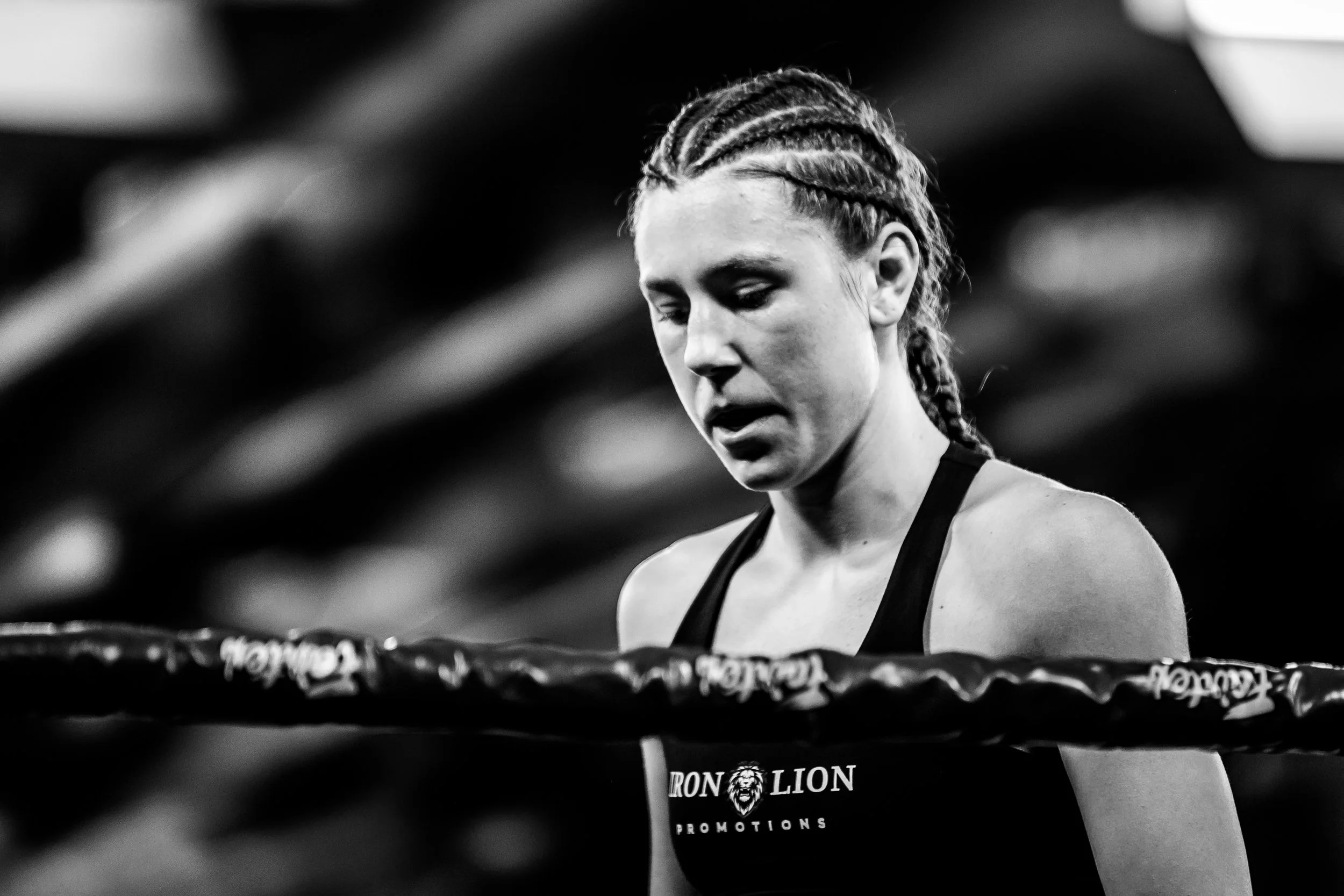 Black and white photo of a female athlete looking down, wearing a tank top with 'Iron Lion Promotions' logo, braided hair, in a gym or workout environment.