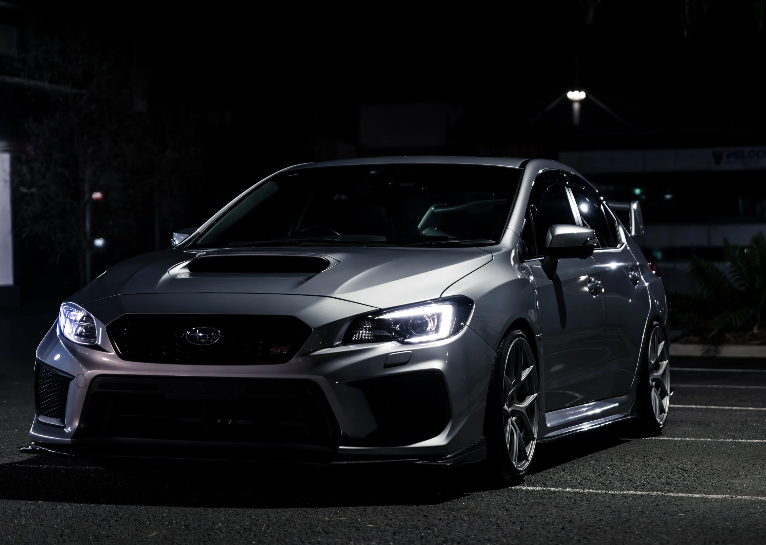 A silver Subaru WRX STI parked at night on an empty parking lot, with headlights on and a dark background.