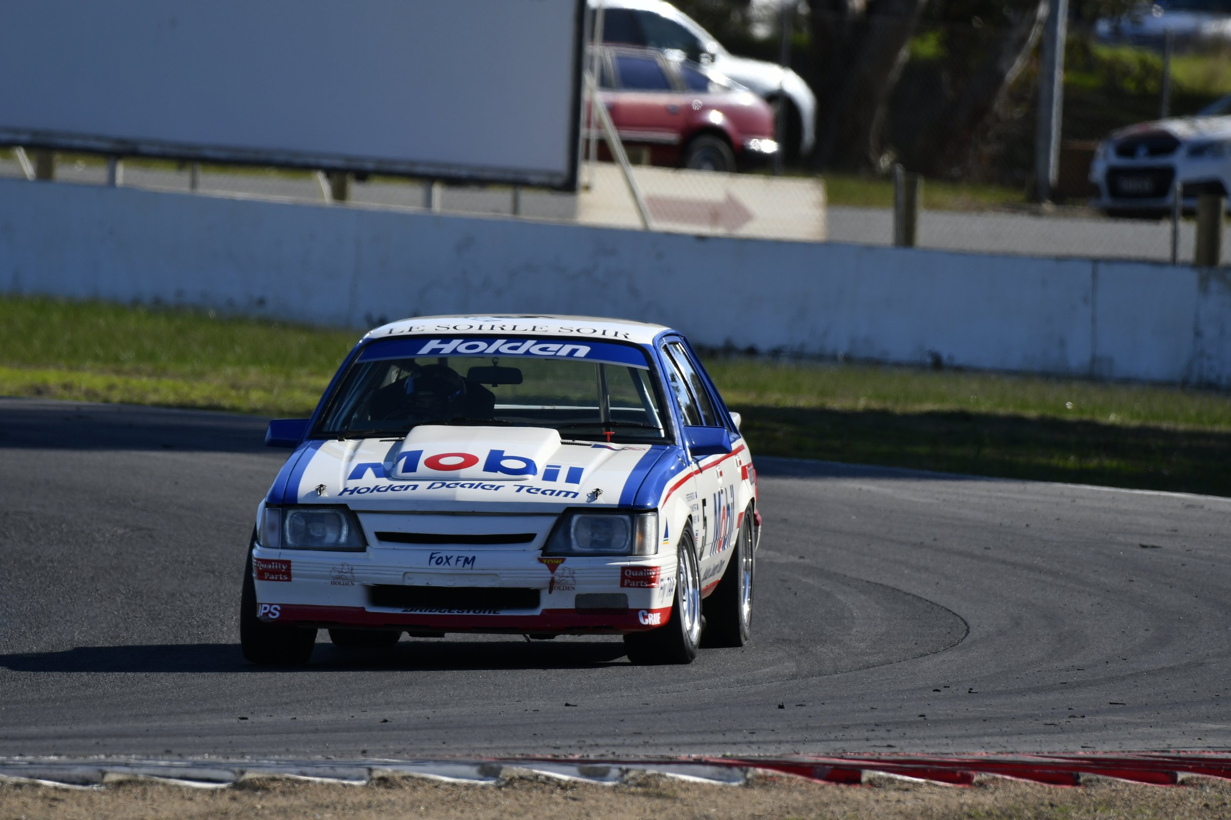 A classic white Holden race car with sponsorship decals, including Mobil, navigating a turn on a race track.