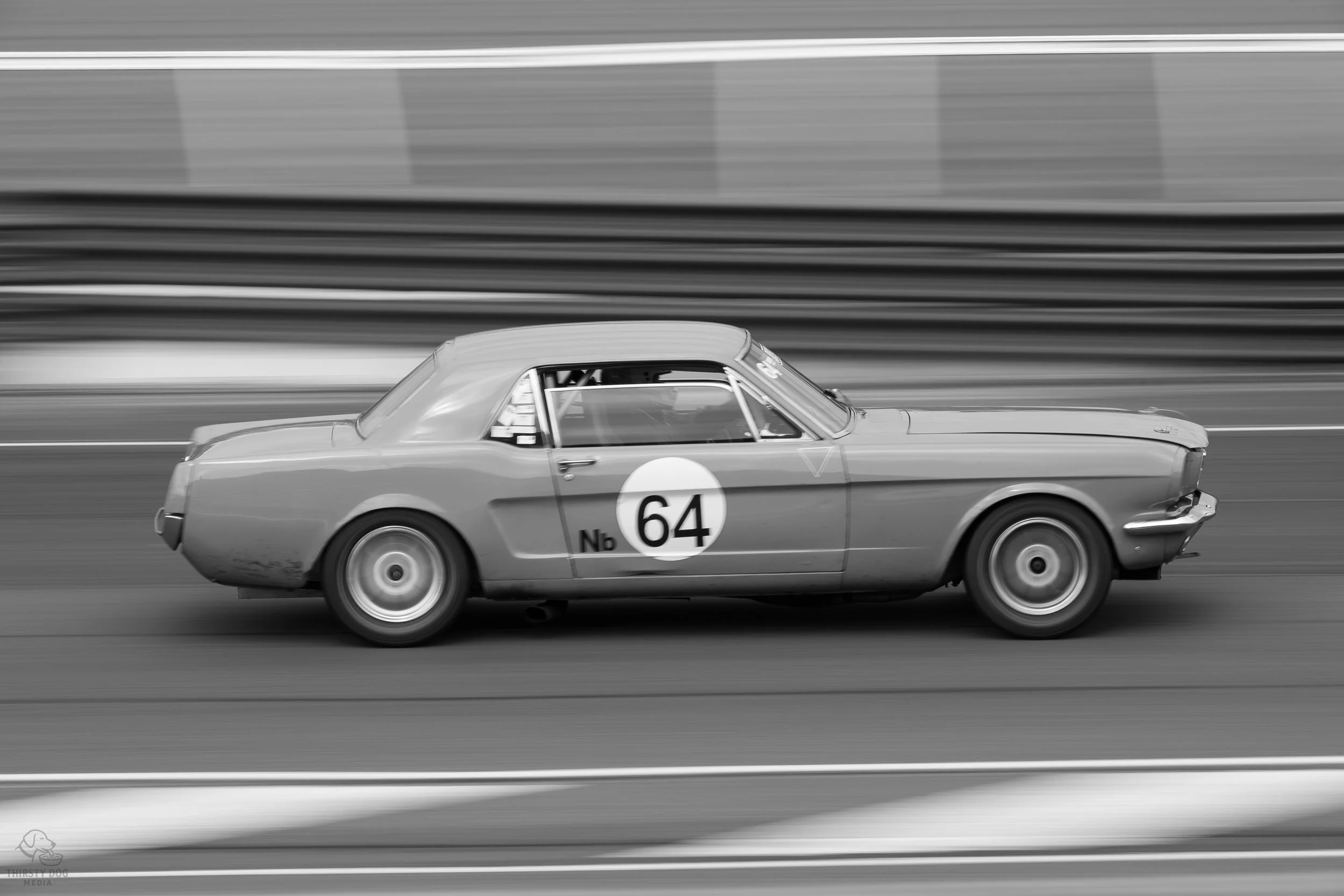 A vintage race car with the number 64 on the door, driving on a racetrack in black and white.