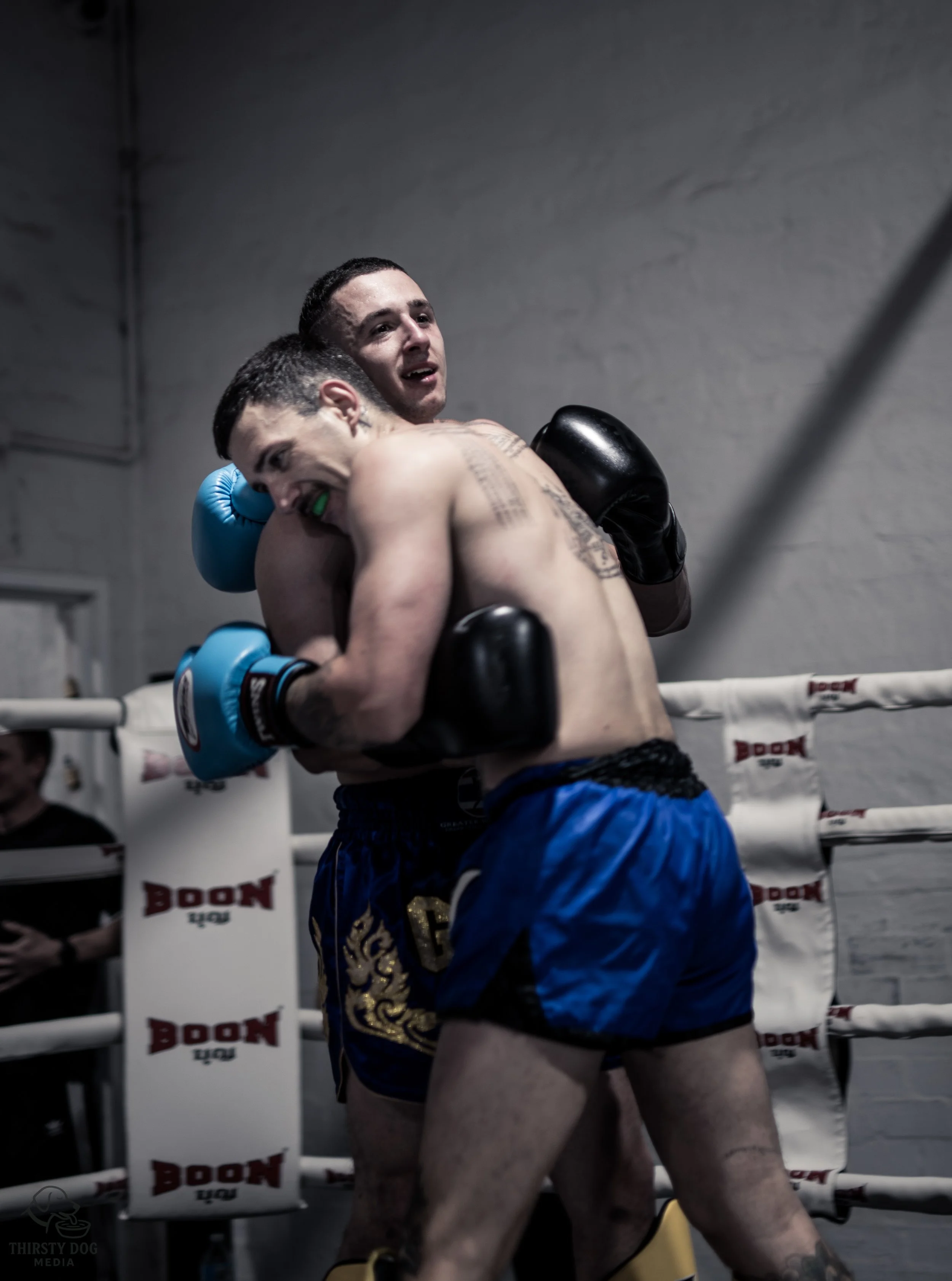 Two male fighters in boxing shorts embracing in the corner of a ring after a match.