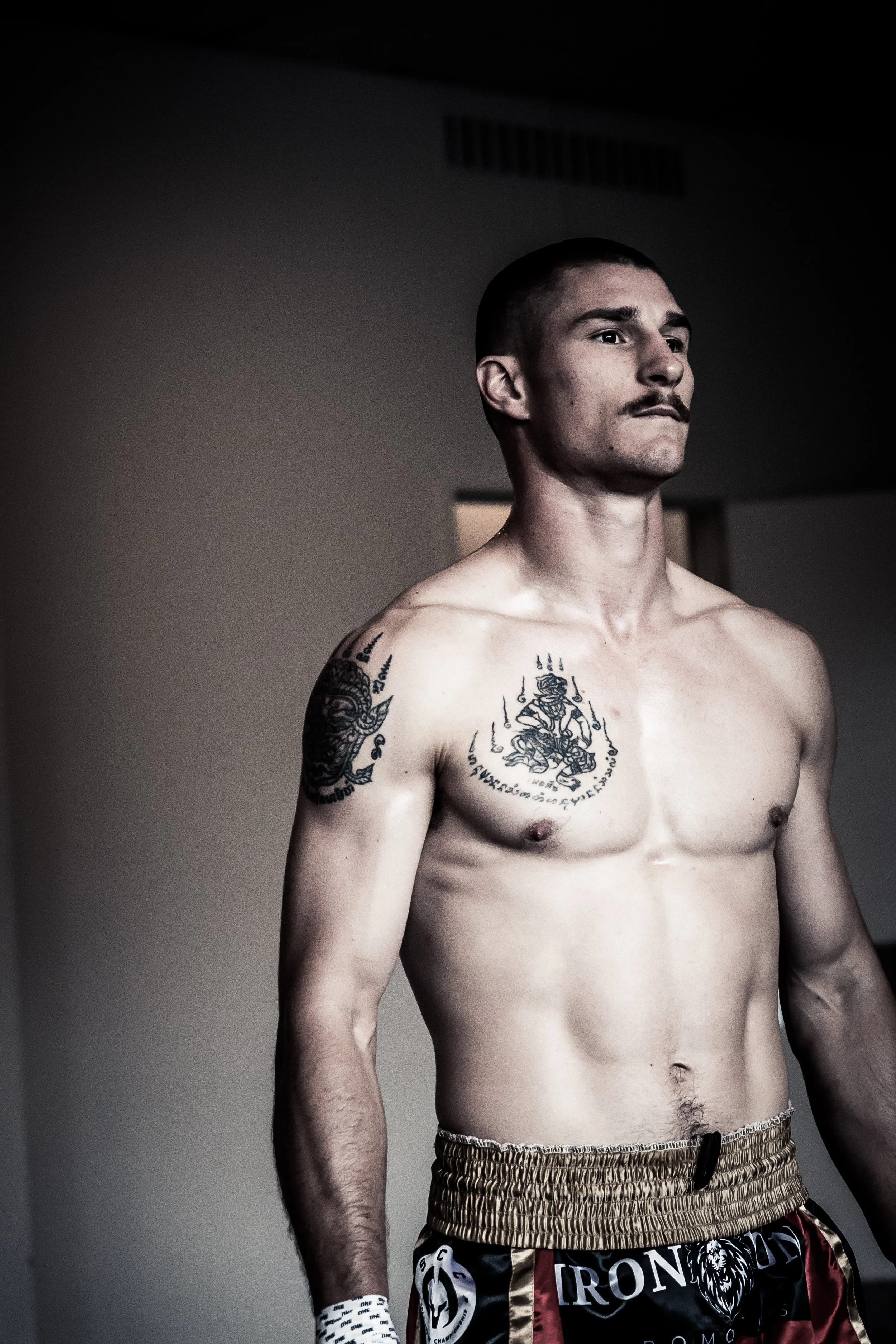 A shirtless man with tattoos on his shoulders and chest, wearing boxing shorts, standing indoors with a serious expression.