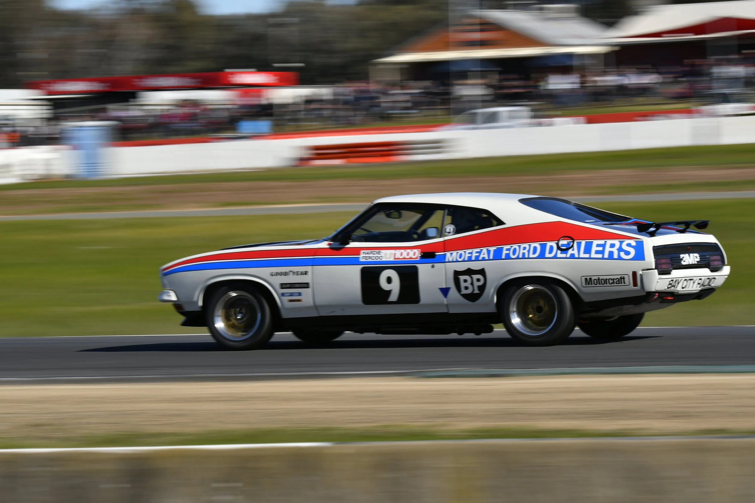 A vintage race car with sponsor logos, including 'Moffat Ford Dealers' and 'BP', speeds along a race track with a blurred background of spectators and race infrastructure.
