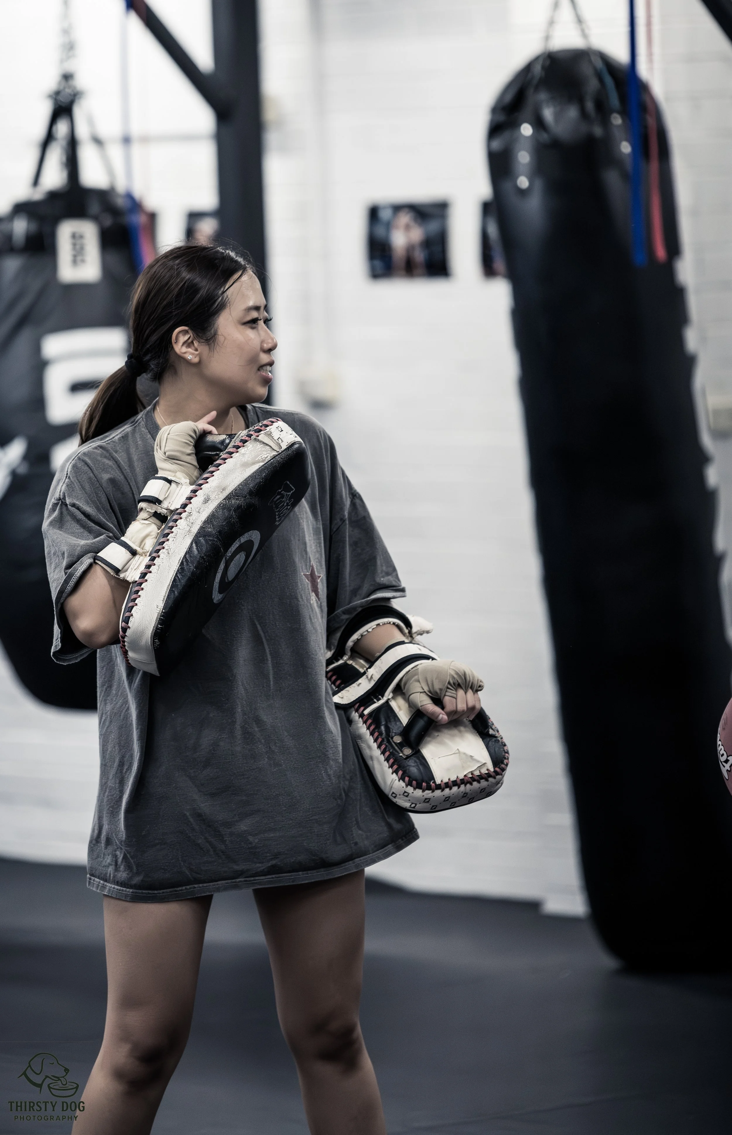 A young woman in a gym holding boxing gloves, standing near punching bags.