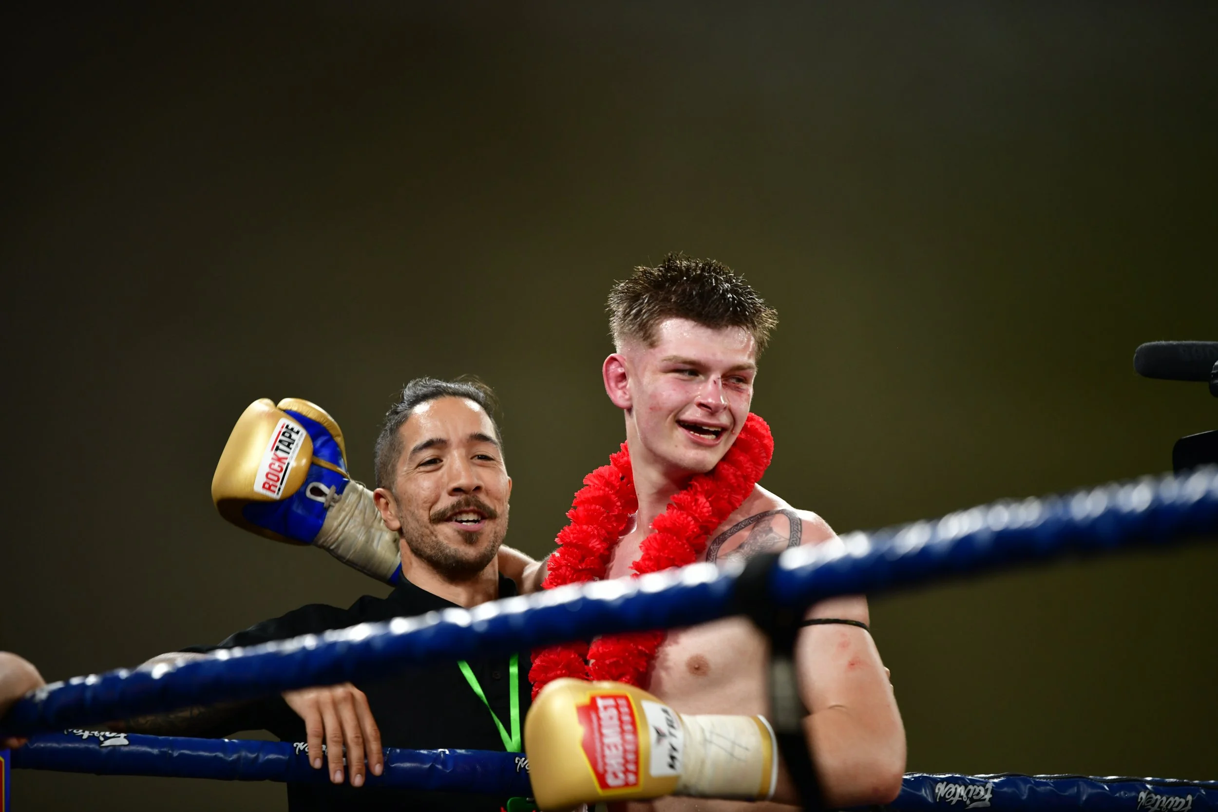 Boxer celebrating with another person, wearing a red lei, in a boxing ring.