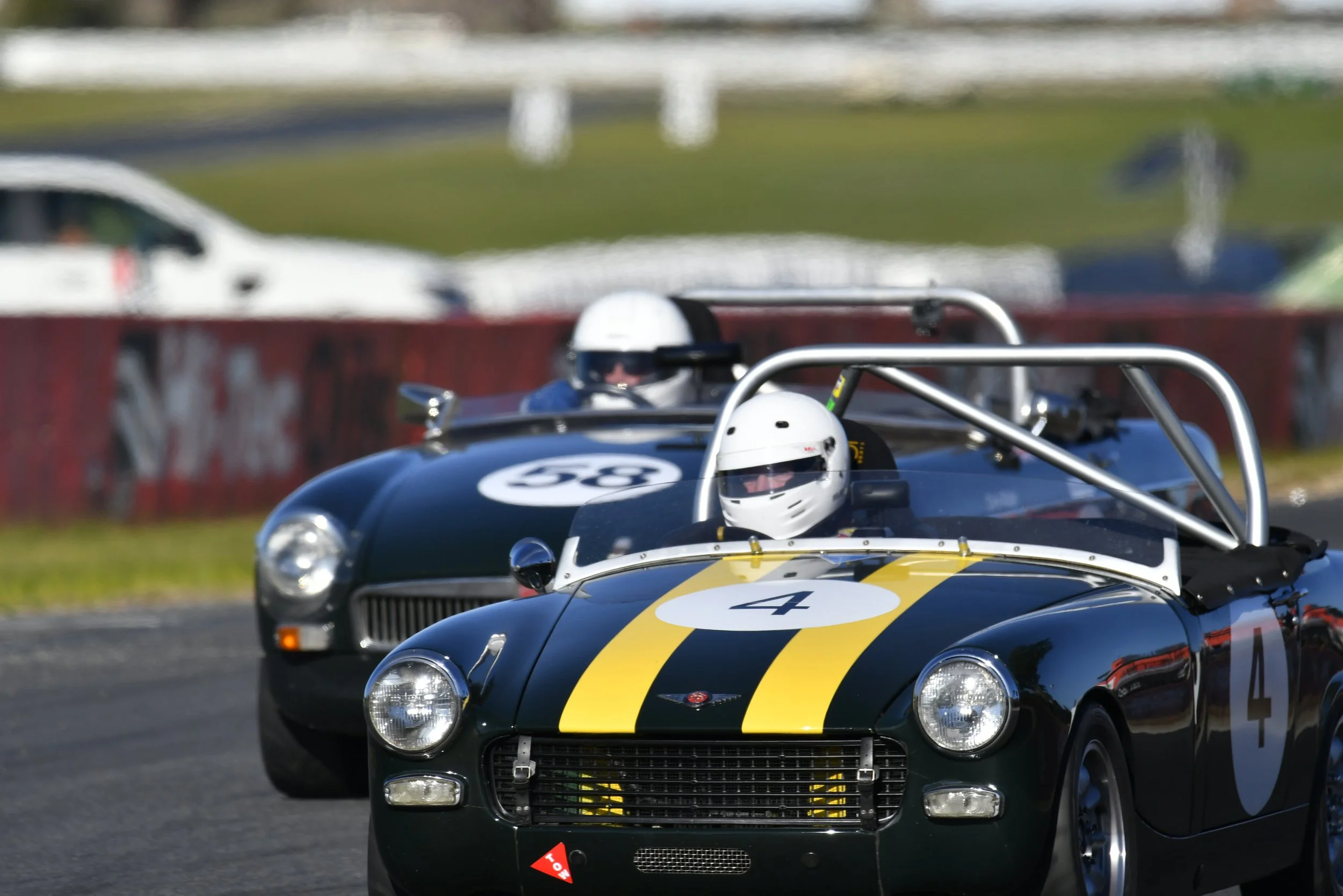 Two vintage race cars with drivers wearing white helmets on a racing track, with a blurred background of cars and grass.