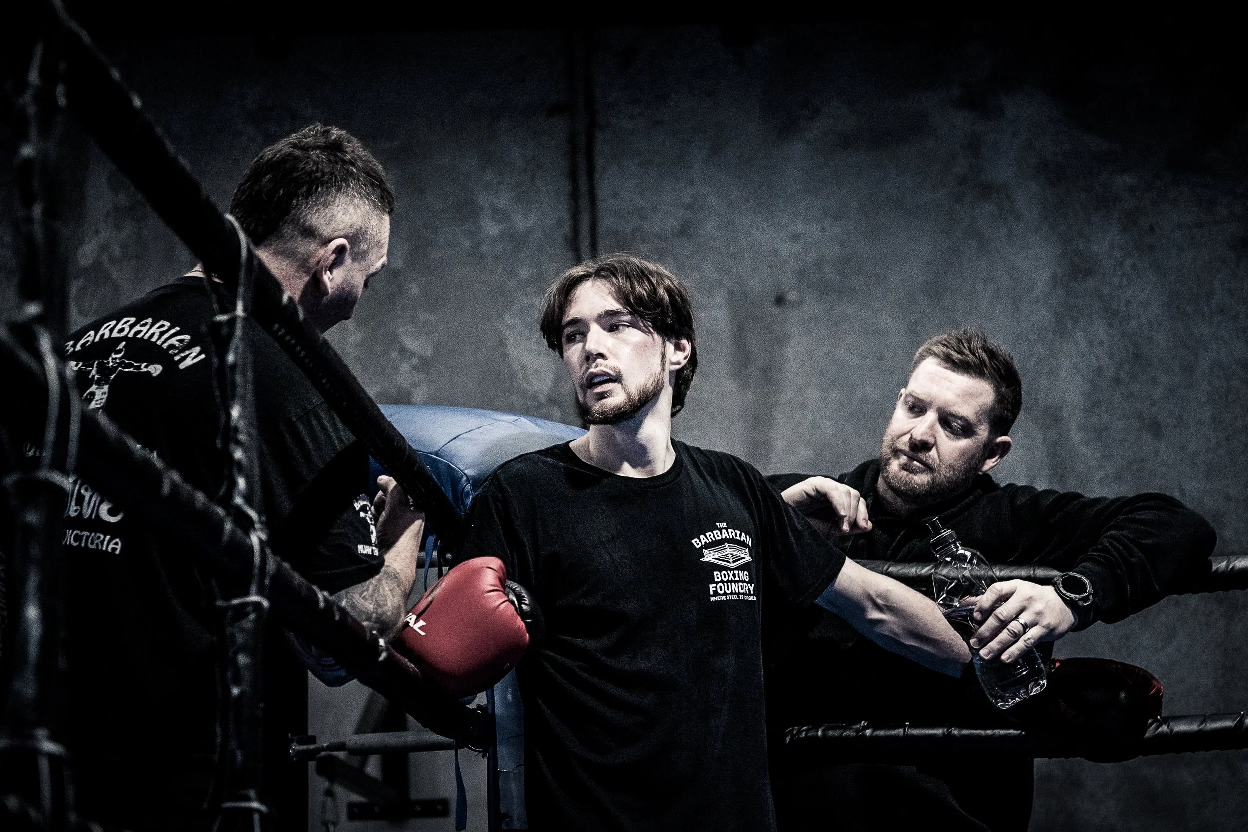 Three men in a boxing gym, one with boxing gloves and two without, engaged in a conversation or coaching session. The man in the middle wears a black shirt with 'The Barbarian Boxing Foundry' logo, while the other two men are listening attentively.