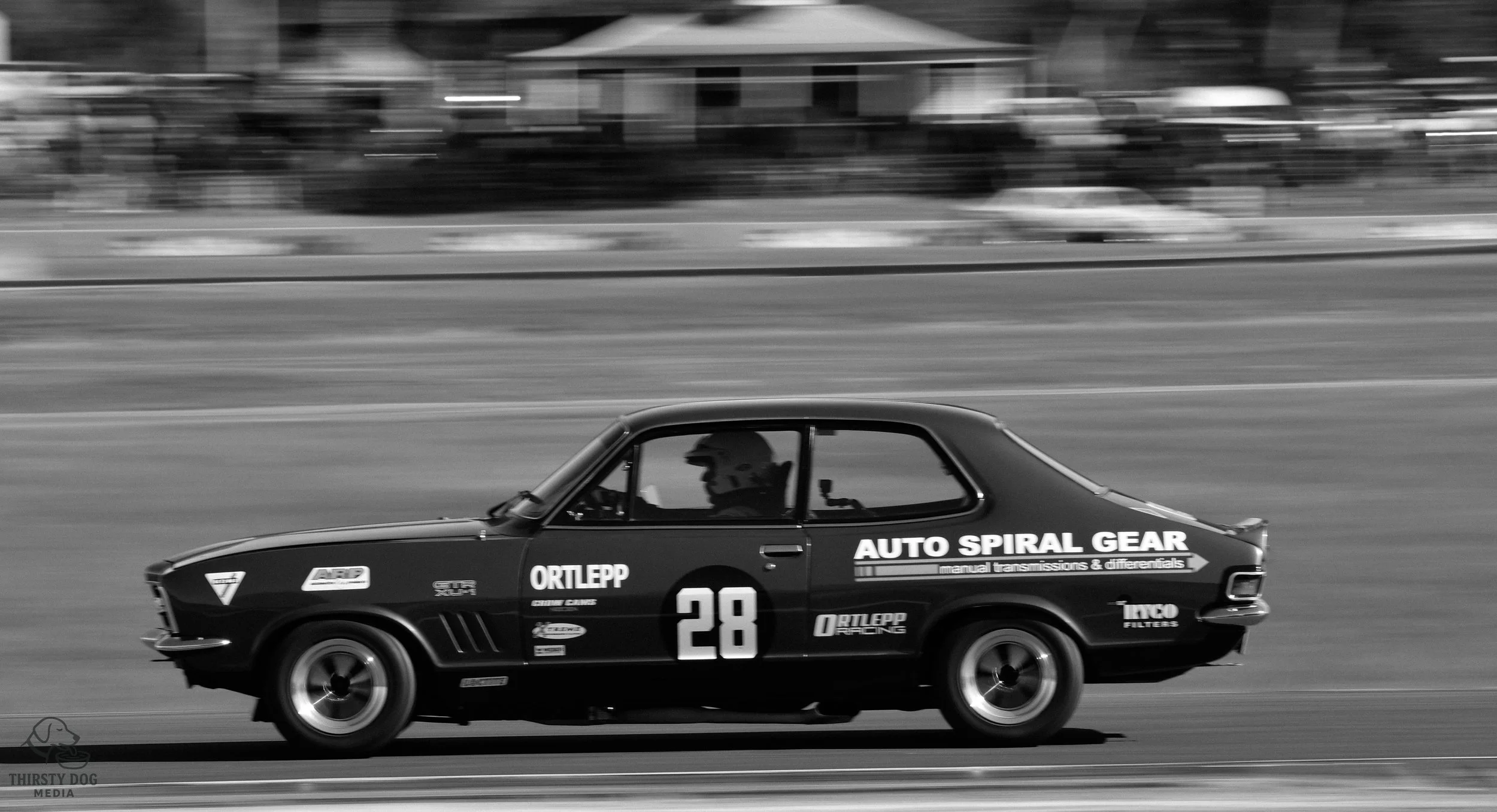 A vintage race car moving at high speed on a race track, with a driver wearing a helmet visible through the side window, in black and white.