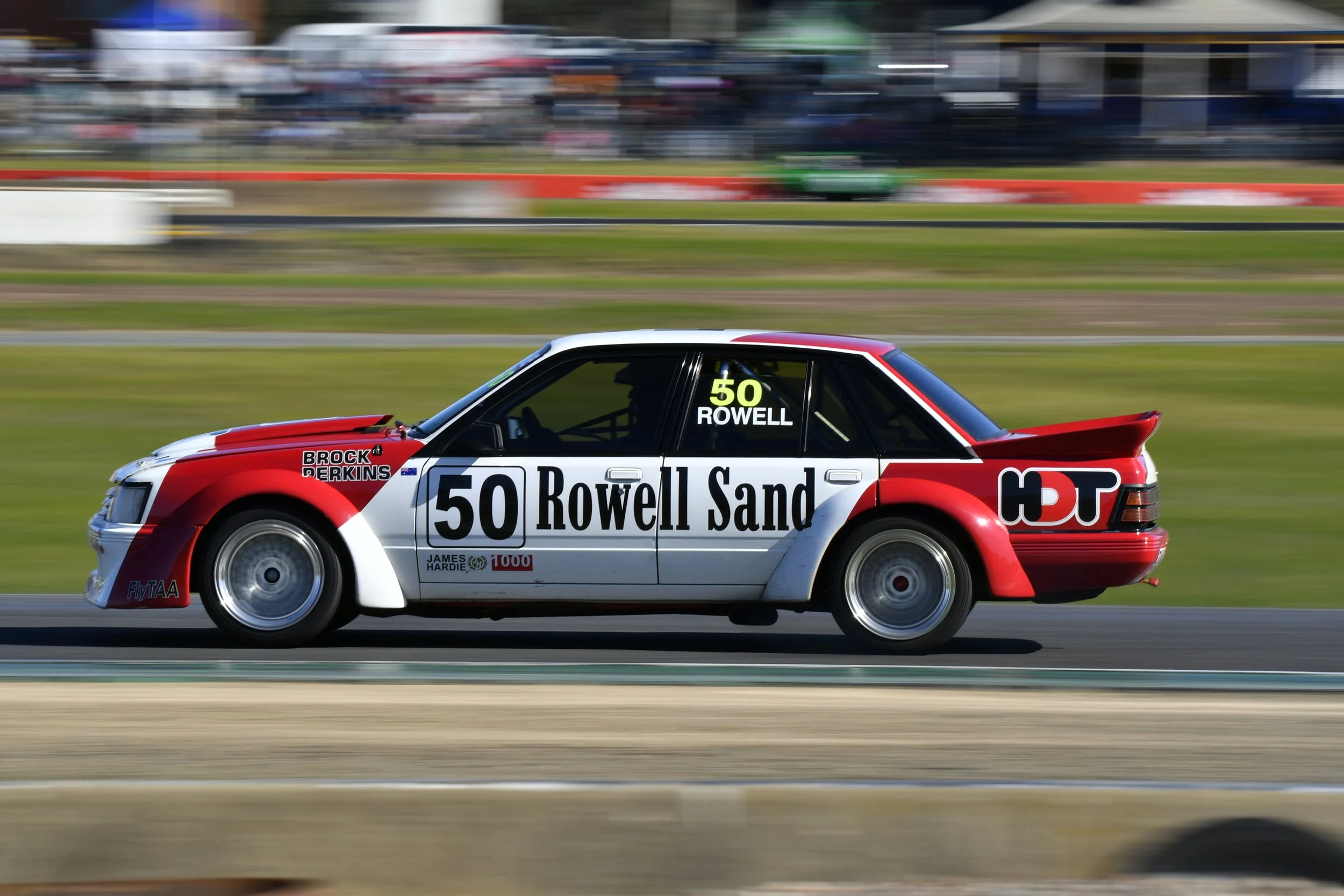A vintage race car with red, white, and black livery moving swiftly on a racetrack, marked with the number 50 and the name Rowell Sand.
