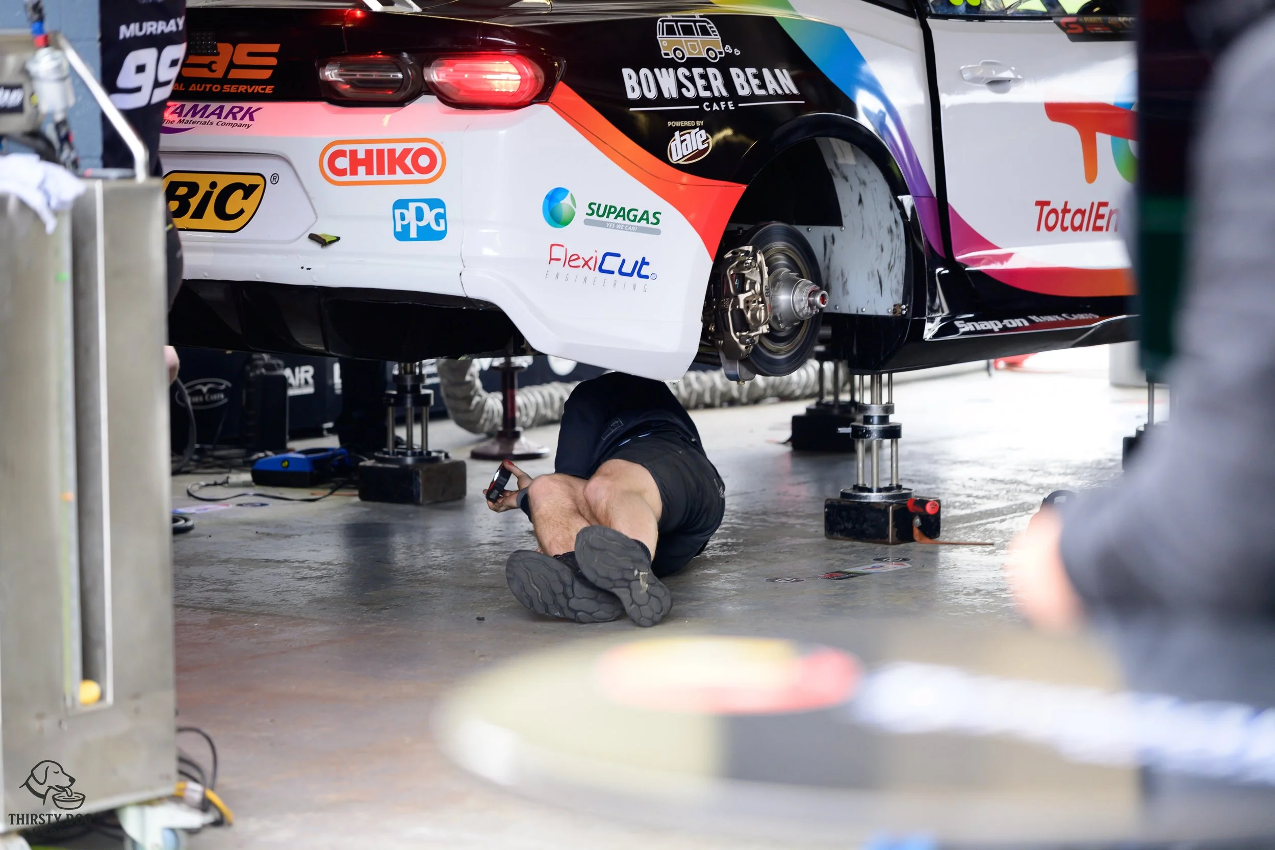 A person working underneath a race car in a garage, with the car elevated on jacks, showing the rear wheel removed and the undercarriage exposed.