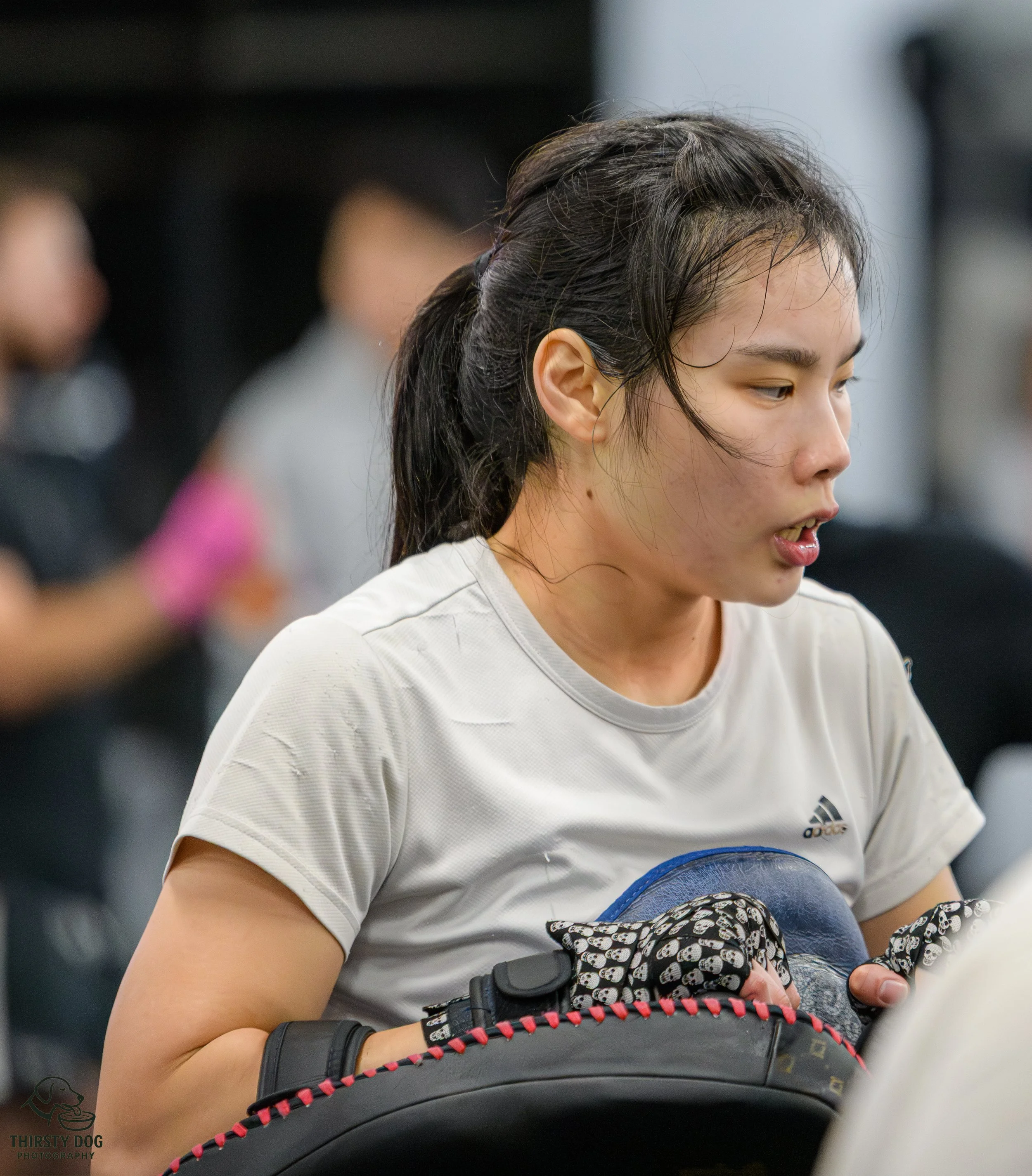 An Asian woman in white sports attire holding a boxing headgear and wearing boxing gloves, appearing tired after a workout or match, with blurred people in the background.