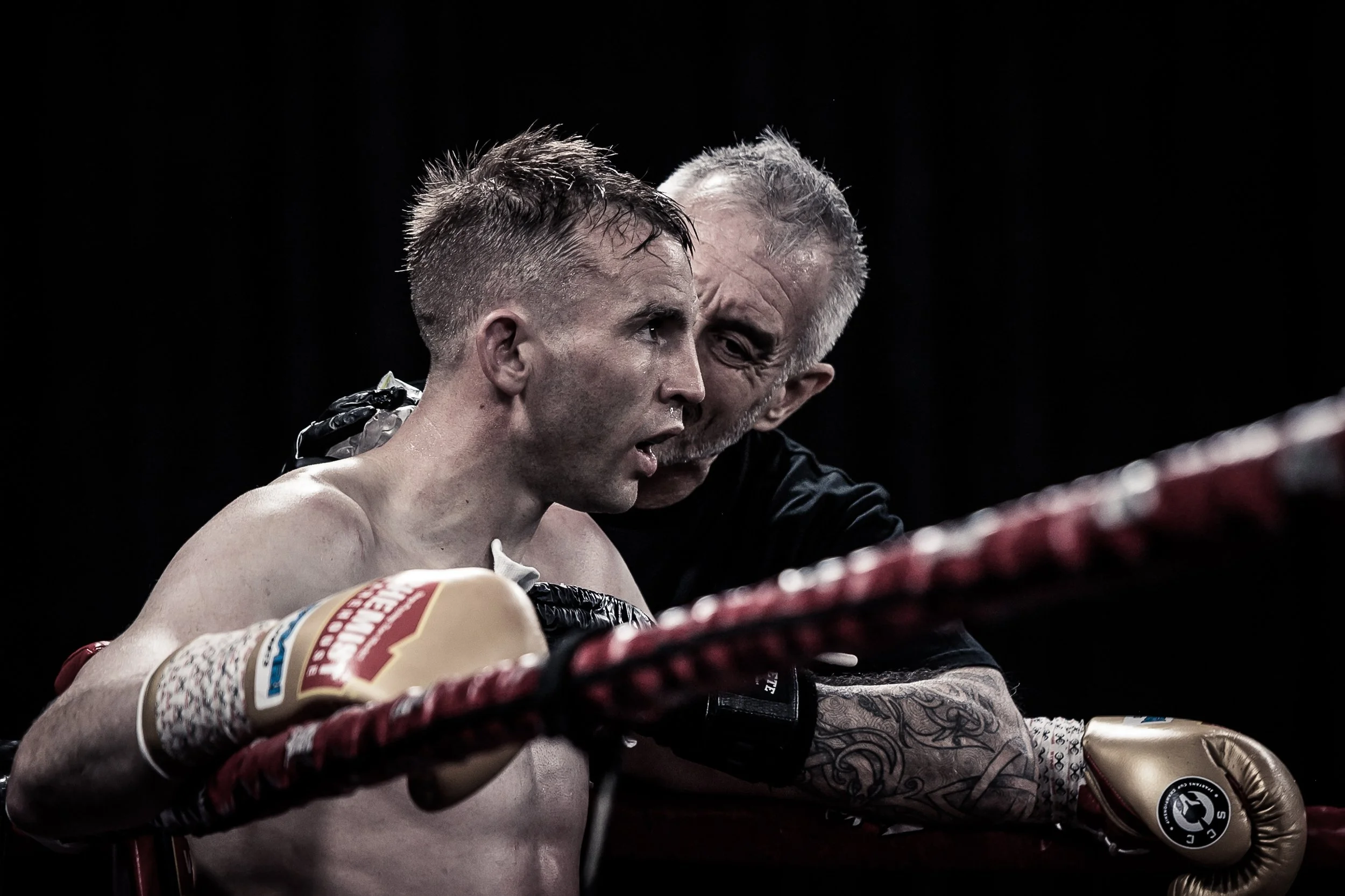 A male boxer sitting in his corner during a match, with his coach leaning over him, giving advice. The boxer is shirtless, wearing boxing gloves and appears to be exhausted and focused.