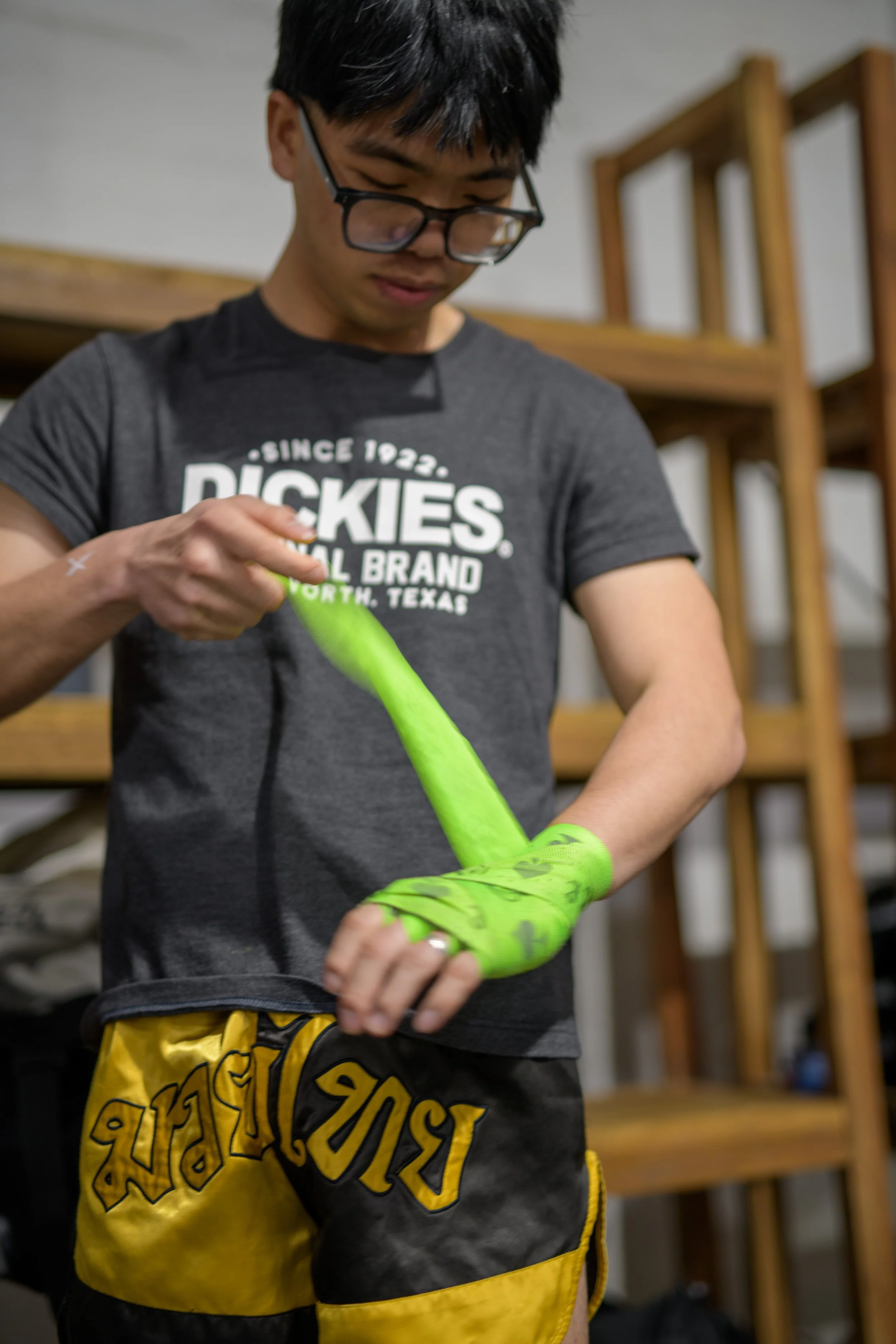 A young man with glasses putting on bright green boxing or sports gloves, standing indoors with wooden furniture in the background.