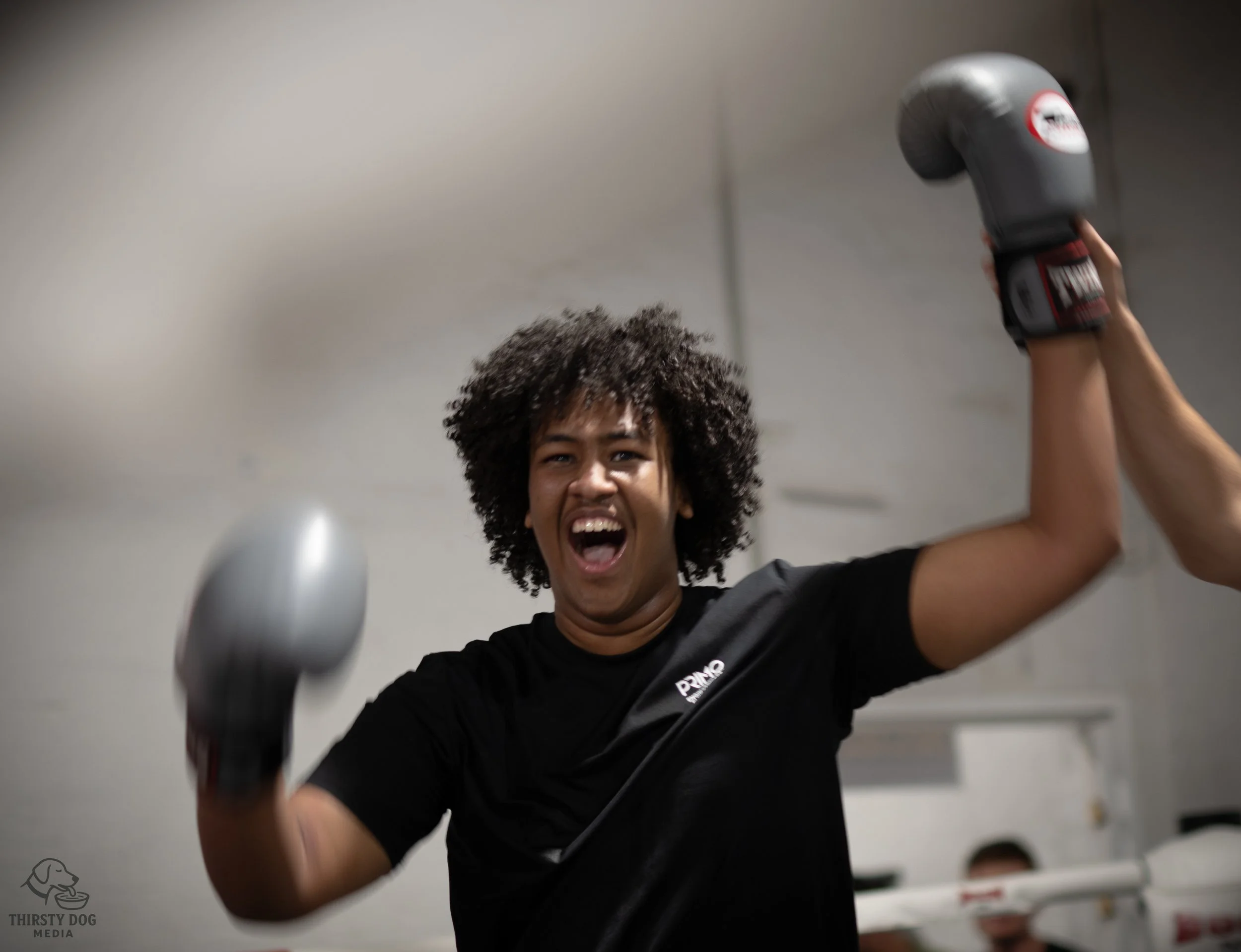 A woman with curly hair wearing a black shirt, smiling and celebrating with boxing gloves in a gym.