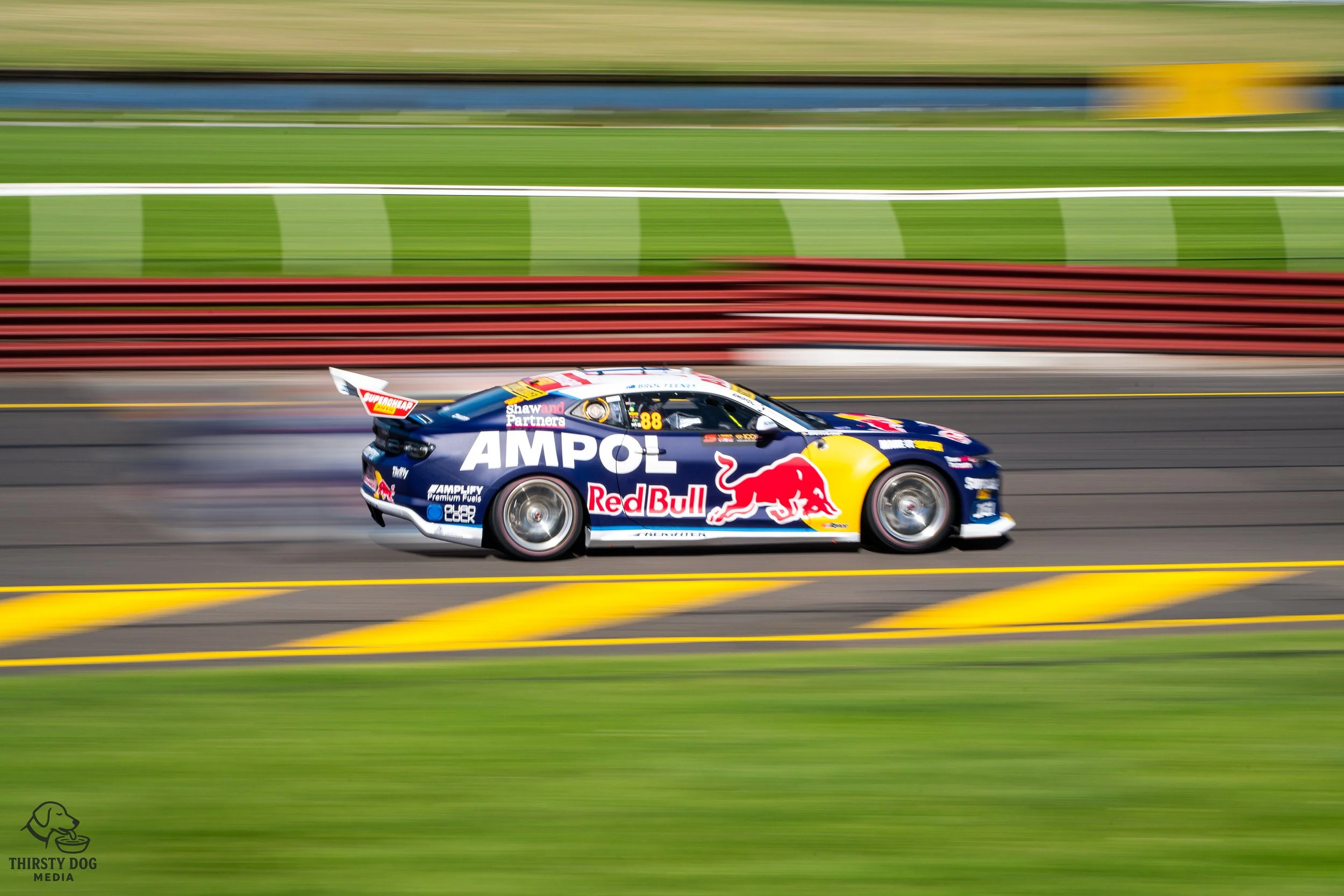 A race car with Red Bull and AMPOL sponsorship logos speeding on a race track with blurred green and red barriers in the background.