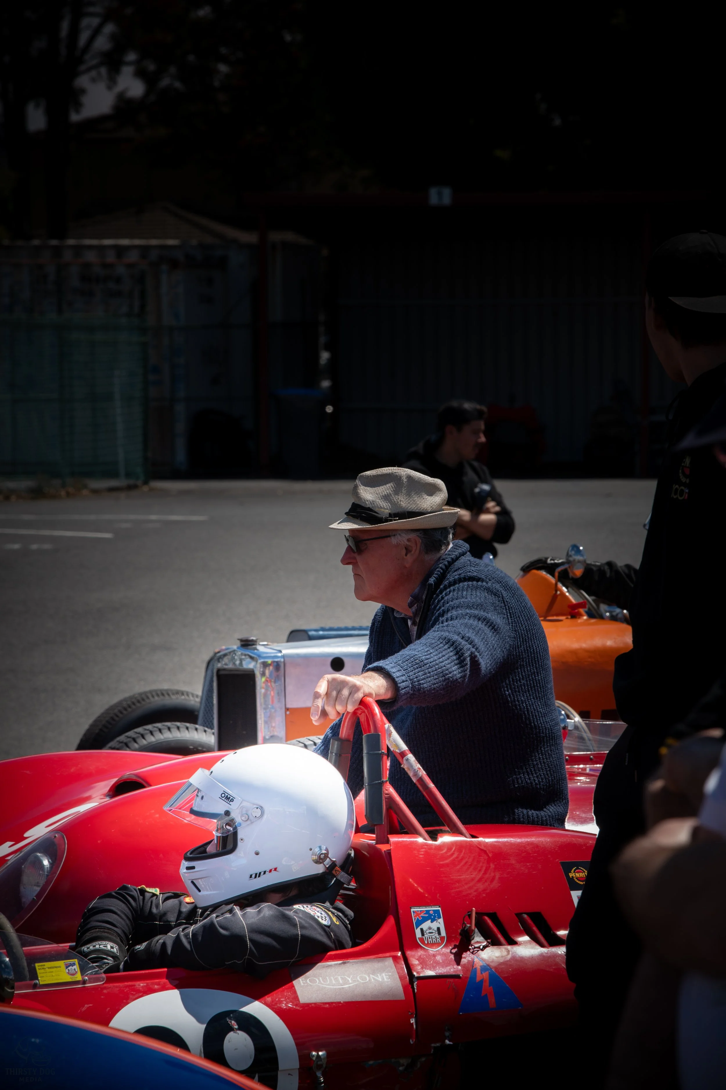 An elderly man in a hat and blue sweater leaning on a red classic race car with a driver wearing a white helmet and racing suit seated inside, while a woman stands in the background and people are gathered around.