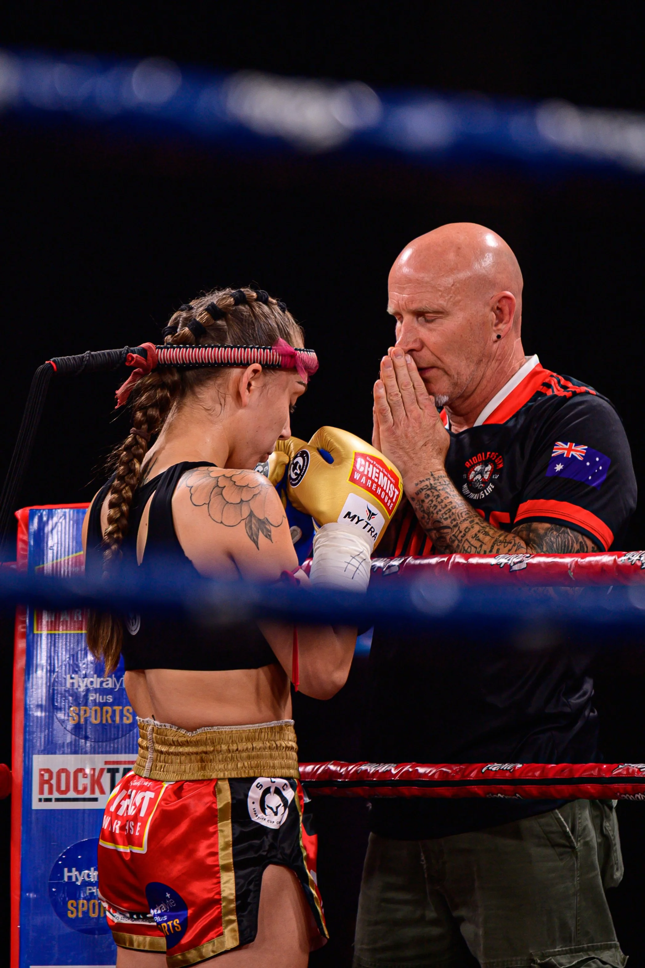A female boxer in boxing gear bows her head with her hands clasped together, holding yellow boxing gloves, while an older male coach with a shaved head and tattoos prays or offers a blessing before a match, inside a boxing ring.