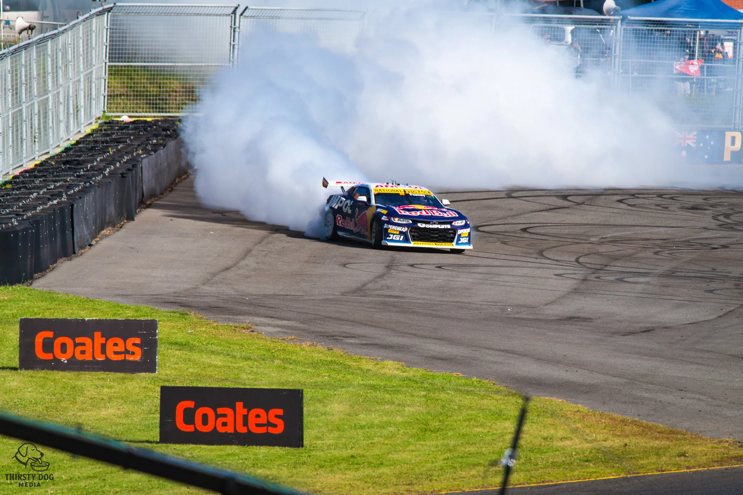A race car performing a burnout on a race track surrounded by tire barriers and smoke. The car has sponsorship logos including Red Bull and FedEx.