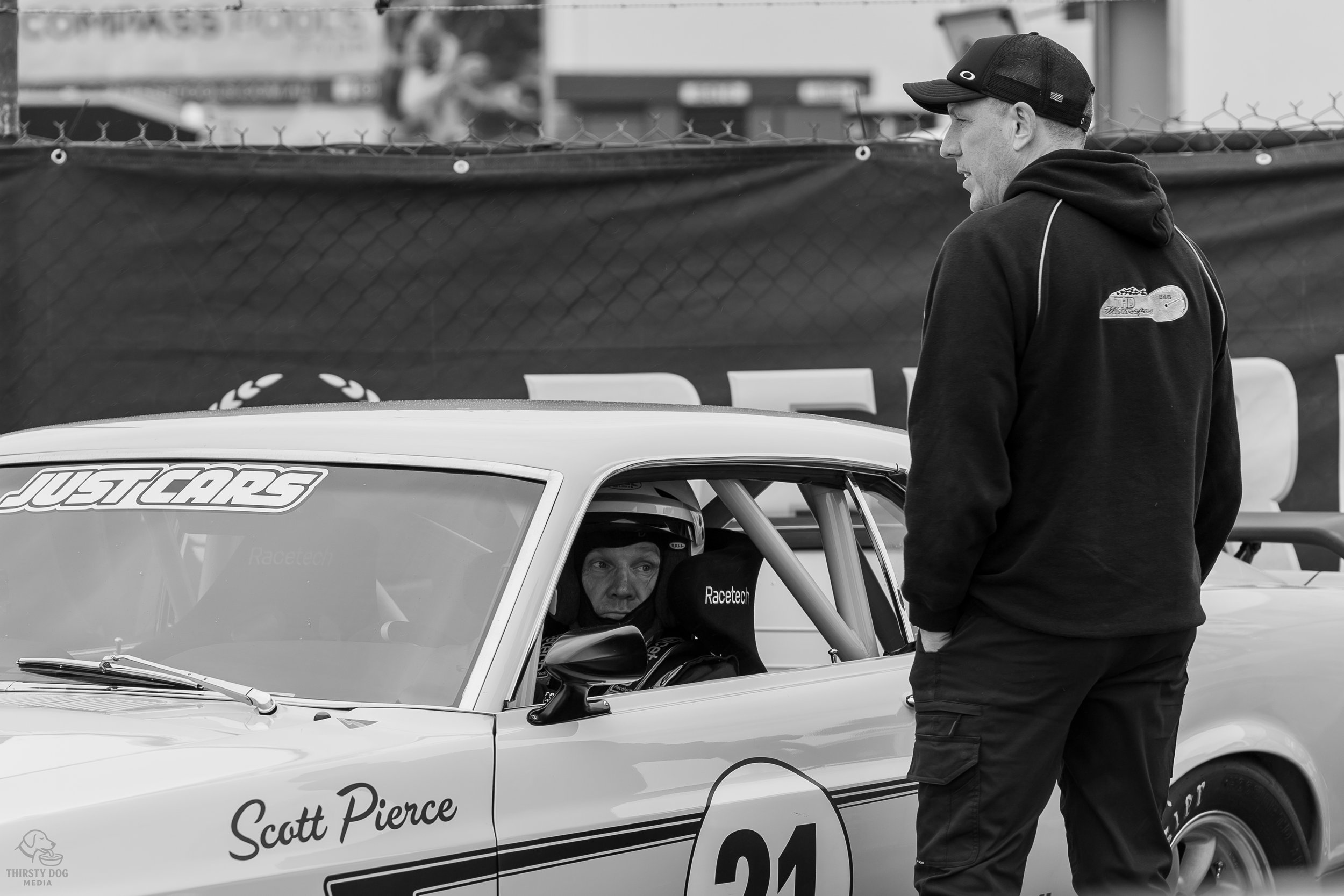 A race car driver wearing a helmet and racing gear sitting inside a vintage race car with the name 'Scott Pierce' on the door, while a man in a black hoodie and cap stands outside. The race car has the number 24 on the side.