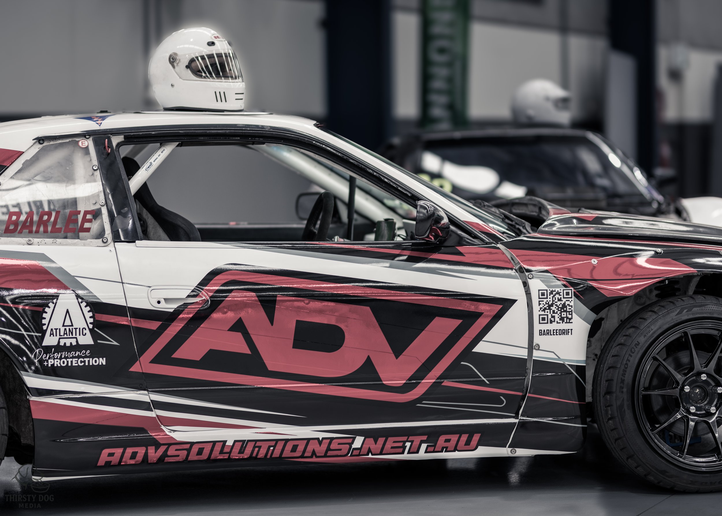 Close-up of a race car with a white helmet placed on top of it, featuring advertising and sponsor logos including 'ADV' and 'ADV SOLUTIONS'. The background shows another vehicle and a garage or pit area.