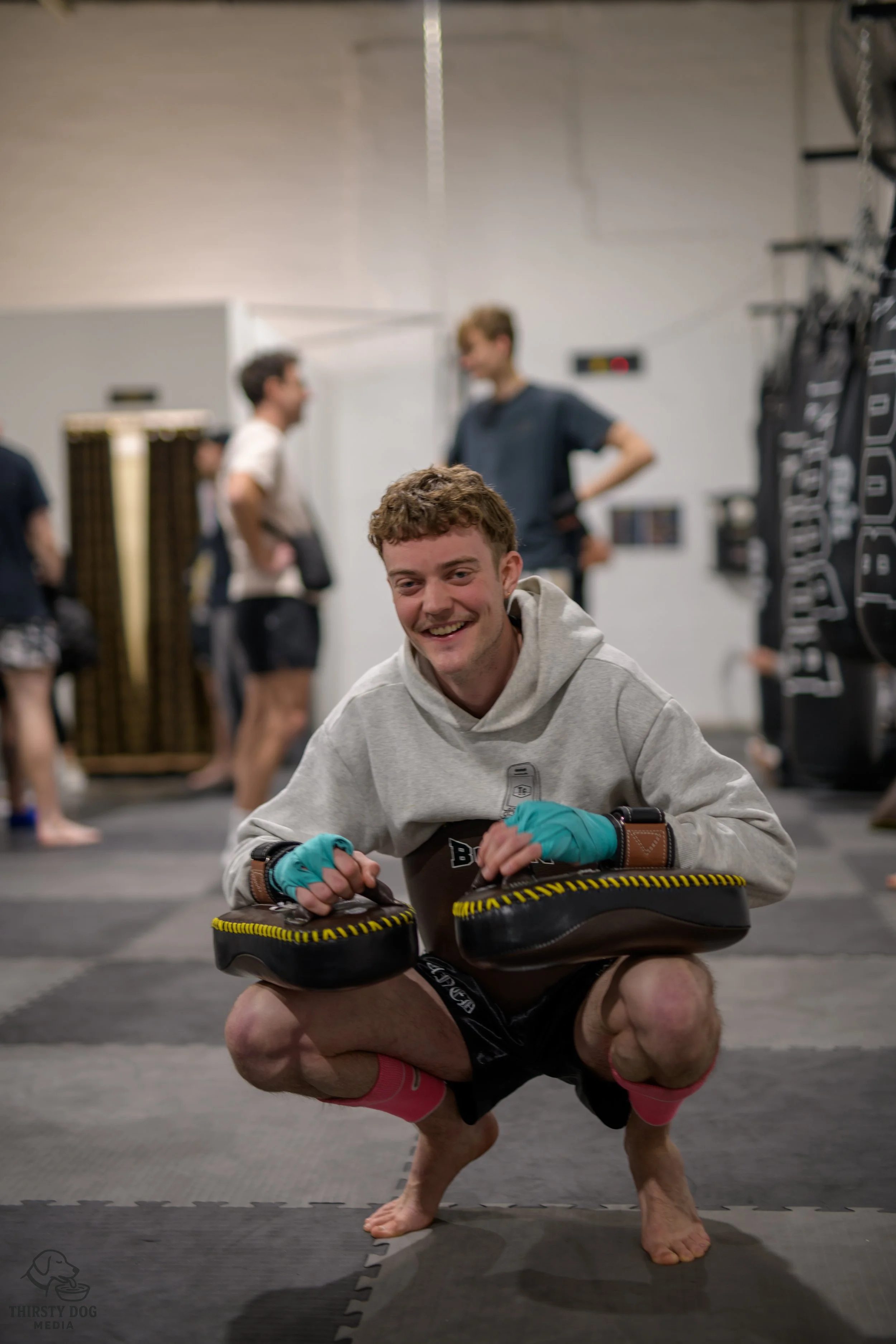 A young man with curly hair smiling, crouching on the floor of a martial arts gym, wearing boxing gloves, a hoodie, and shorts, with others in the background.
