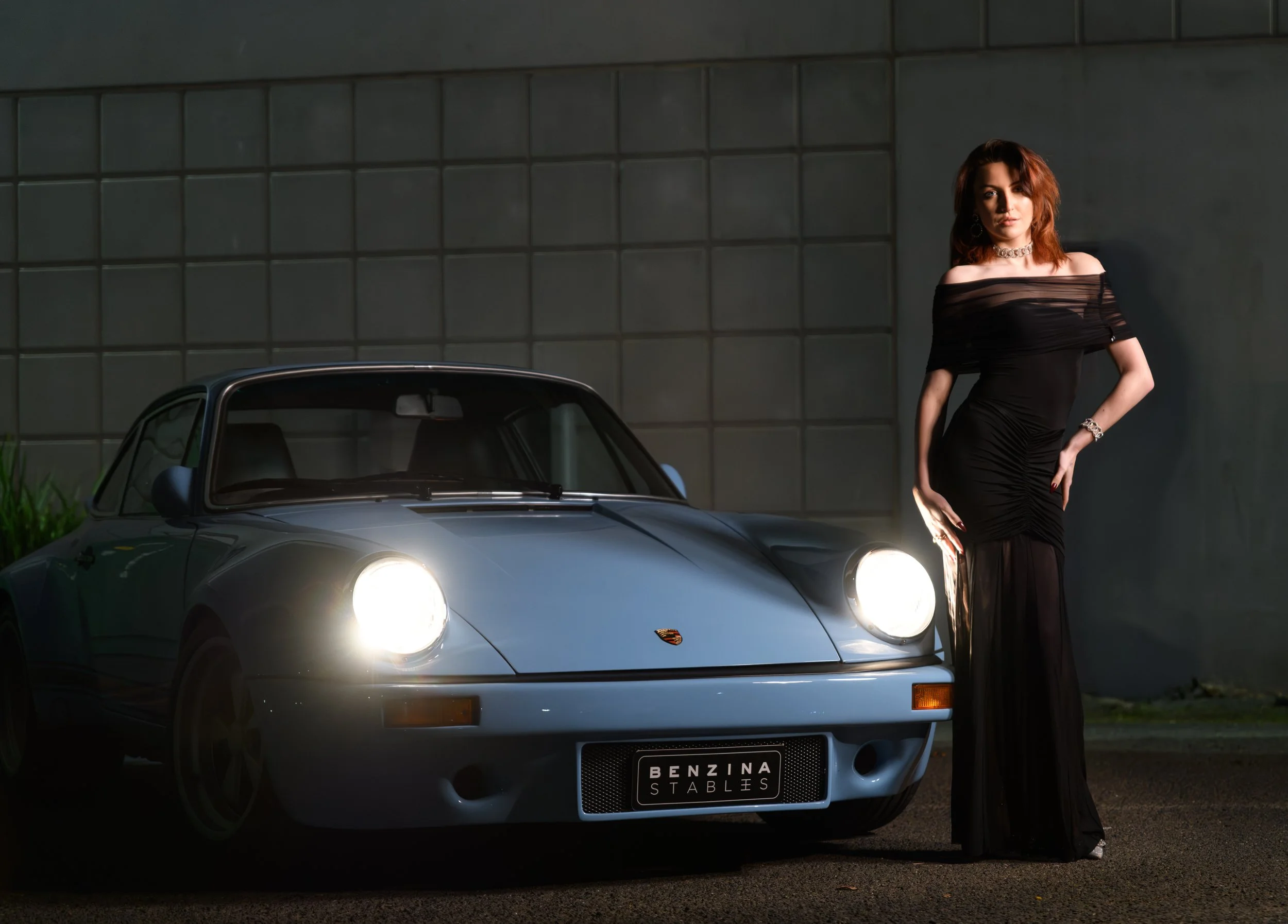 A woman in a black evening gown standing next to a classic blue Porsche sports car outdoors at night.