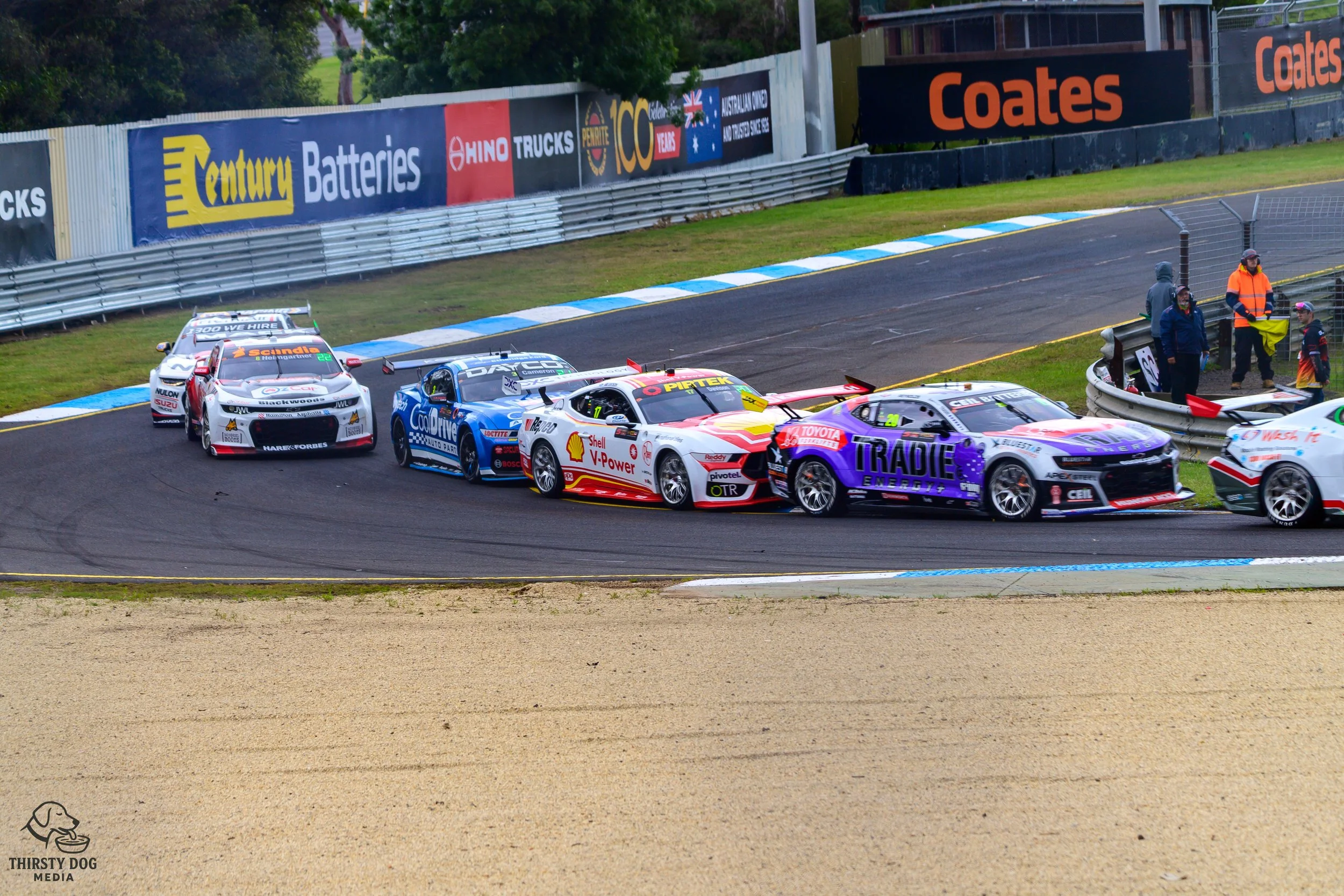 Multiple race cars collide on a racetrack, with some cars against the guardrail and others in the middle of the track. Several people and officials are watching from the sidelines.