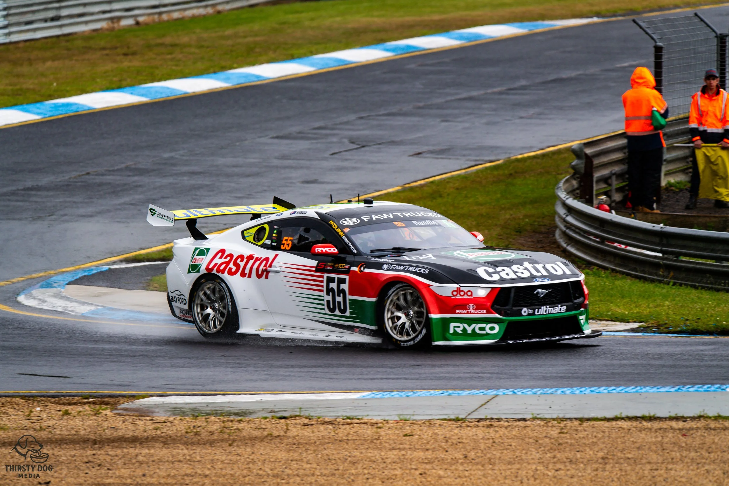 Race car driving on wet racing track with two officials in orange safety vests standing near safety barrier in background.