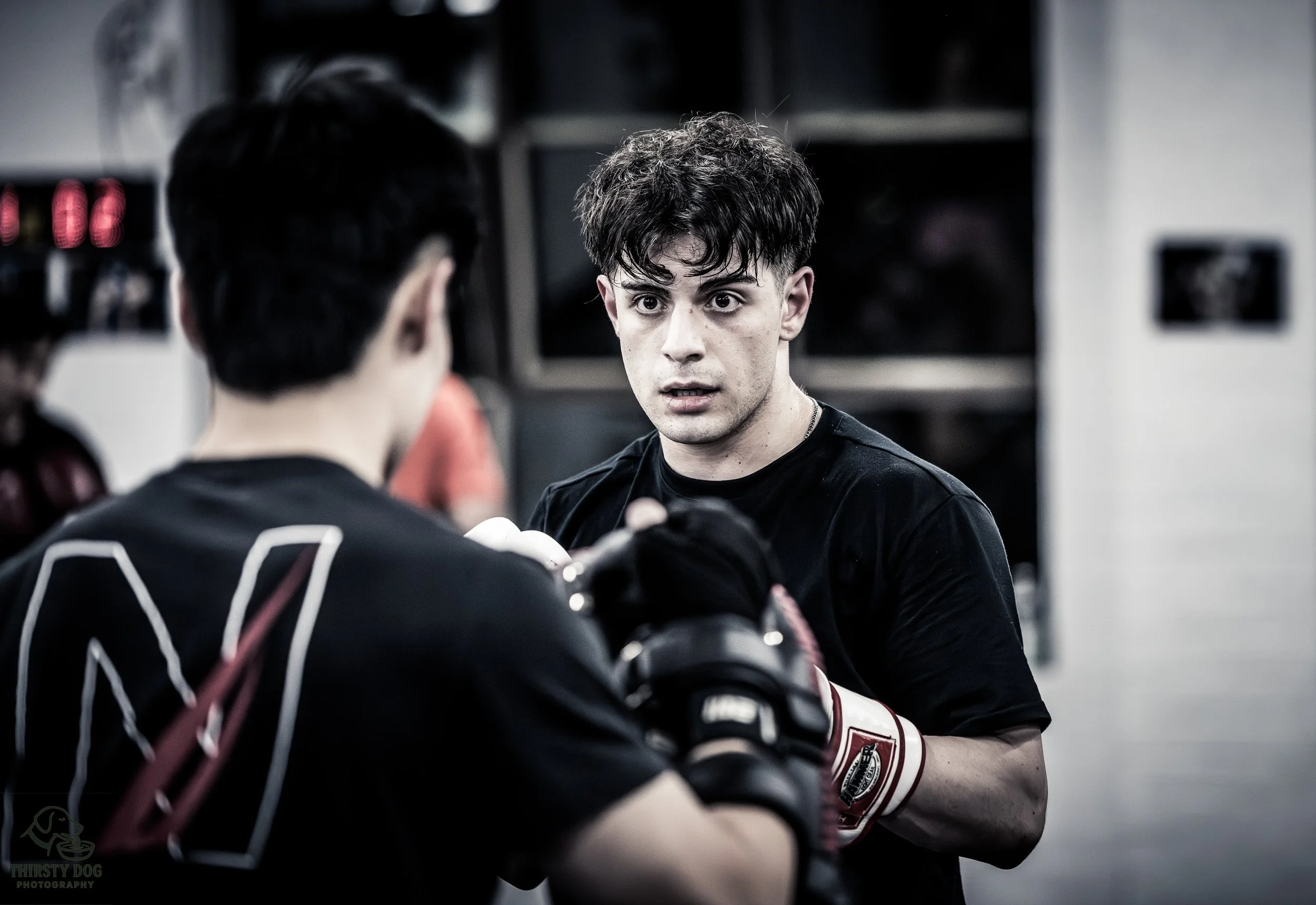 Two young men boxing in a gym, with one wearing boxing gloves, appearing focused during training.
