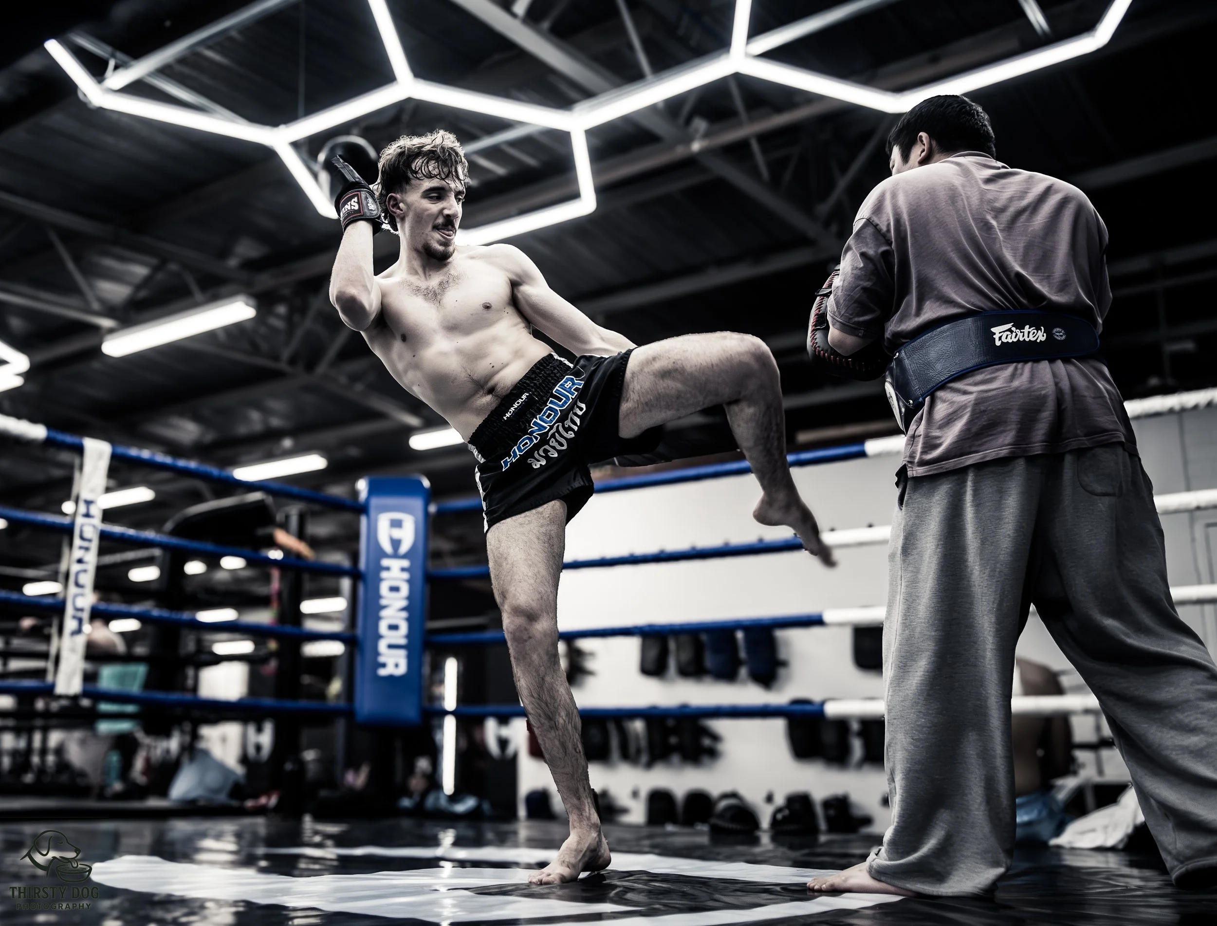 A shirtless male mixed martial artist practicing strikes with a coach in a gym boxing ring. The fighter is in mid-motion, raising his knee, wearing black shorts, with gloves on. The coach faces him, wearing a gray shirt and pants, holding focus mitts