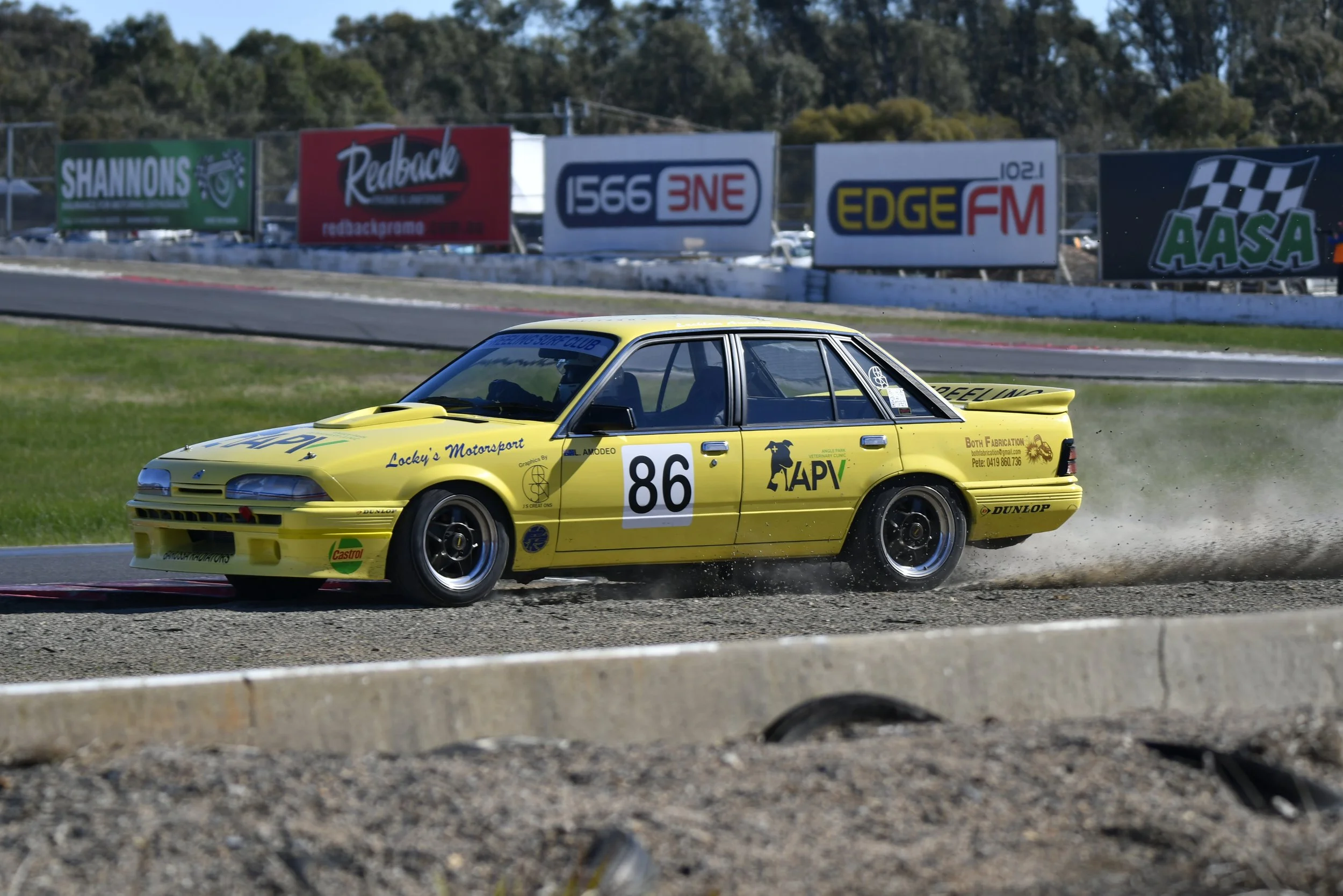 A yellow race car with the number 86 on the side driving on a dirt track, kicking up dust behind it, with advertisements on billboards in the background.