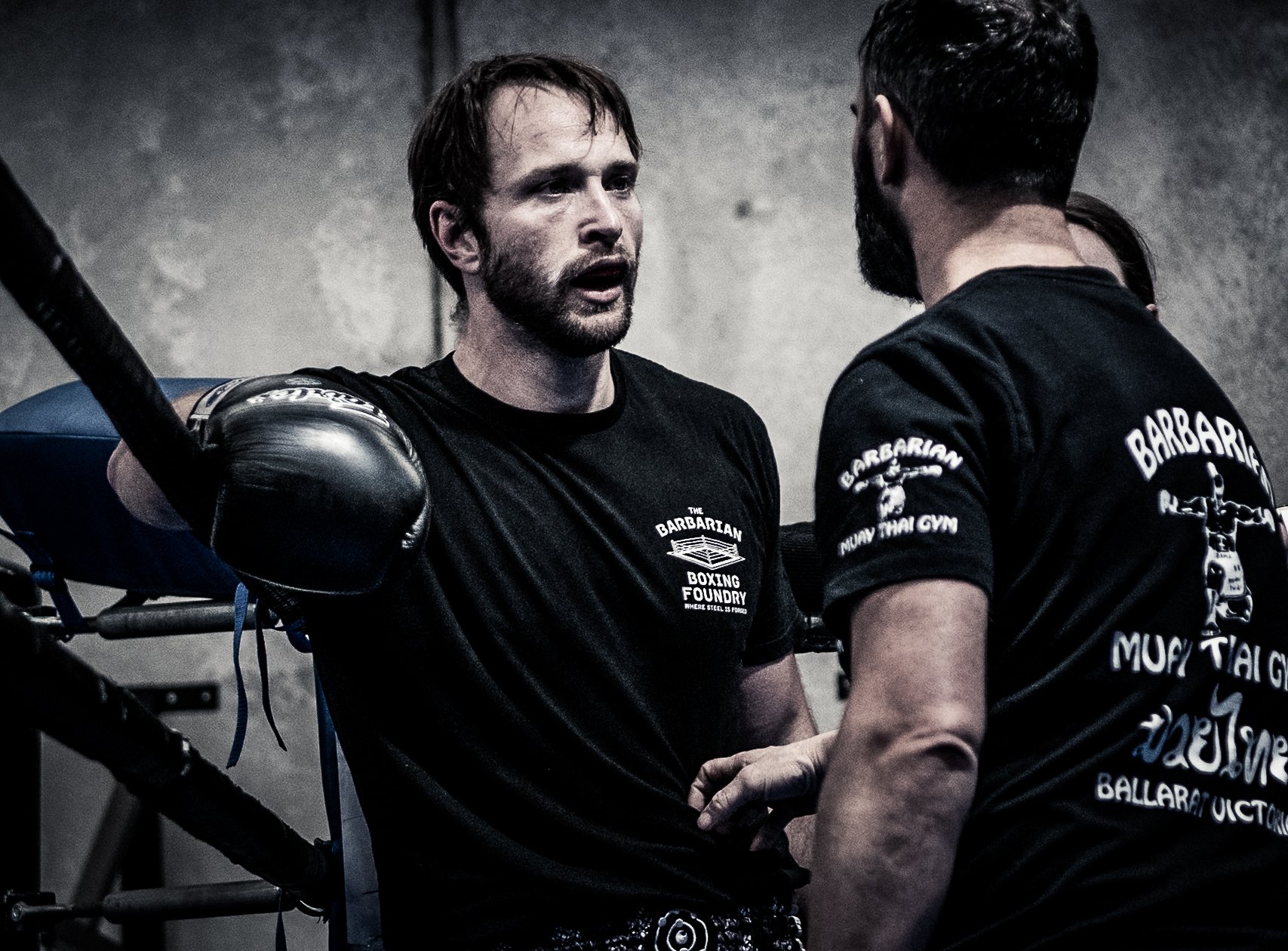 Two men in black t-shirts with martial arts logos having a serious conversation in a gym. One man is wearing boxing gloves and leaning on a punching bag, while the other stands close, engaging in the discussion.