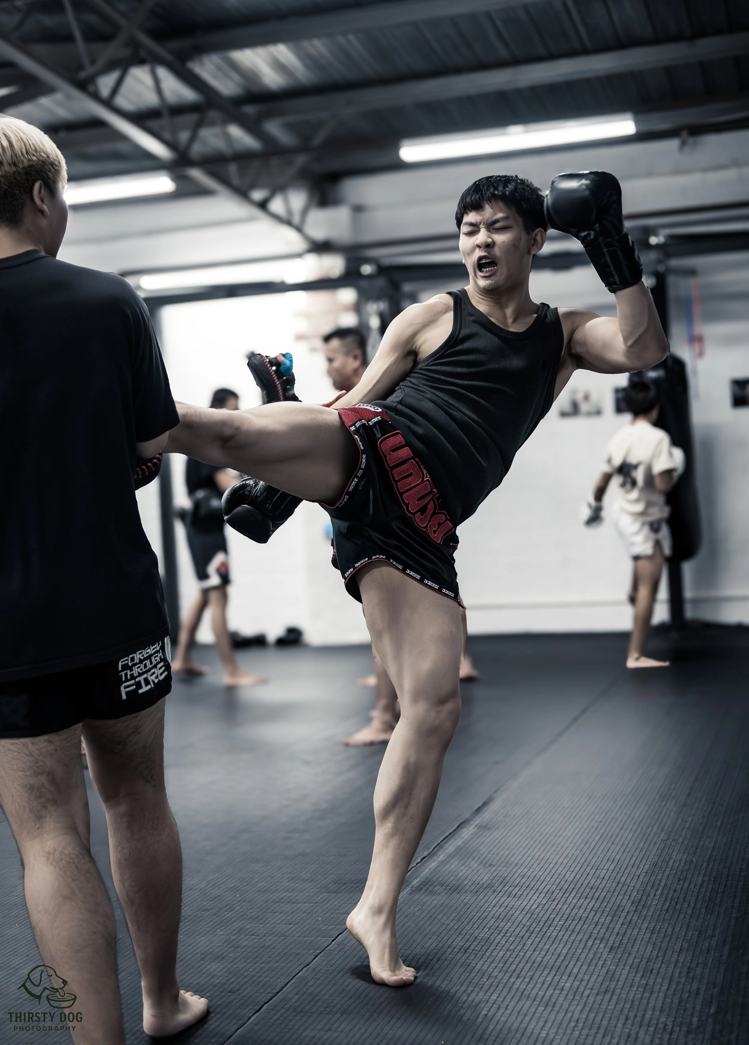 A young man practicing kickboxing at a gym with other people in the background. He is executing a high kick towards a partner who is holding his shin. The martial artist wears black shorts and a black tank top, with gloves on both hands.