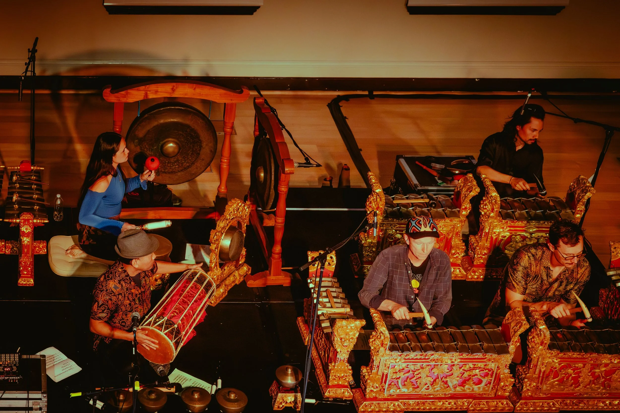 Musicians playing traditional instruments on stage, including a woman with a gong, a man with a drum, and others with metallophones, in an ornate setting.