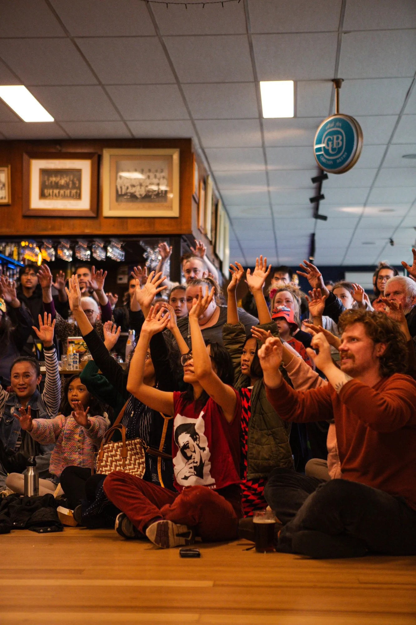 Crowd of people sitting and standing in a bar, raising their hands during an event or performance, with framed pictures on the wall and a round sign hanging from the ceiling.