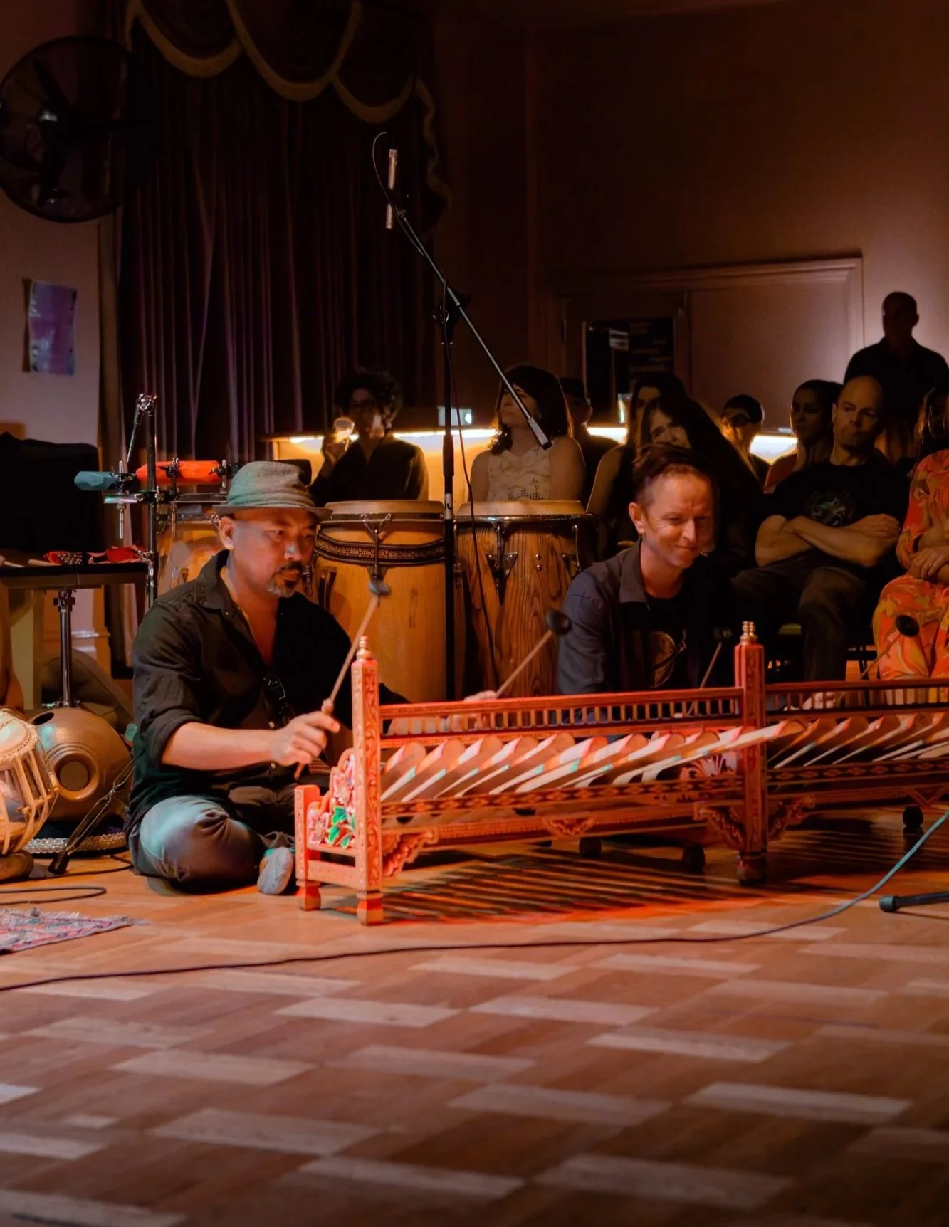Two musicians sit on the floor playing traditional drums and a stringed instrument in a dimly lit venue with an audience watching.