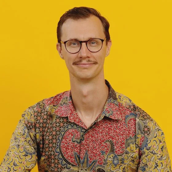 Portrait of a man with glasses and a mustache, wearing a colorful patterned shirt, standing against a yellow background.