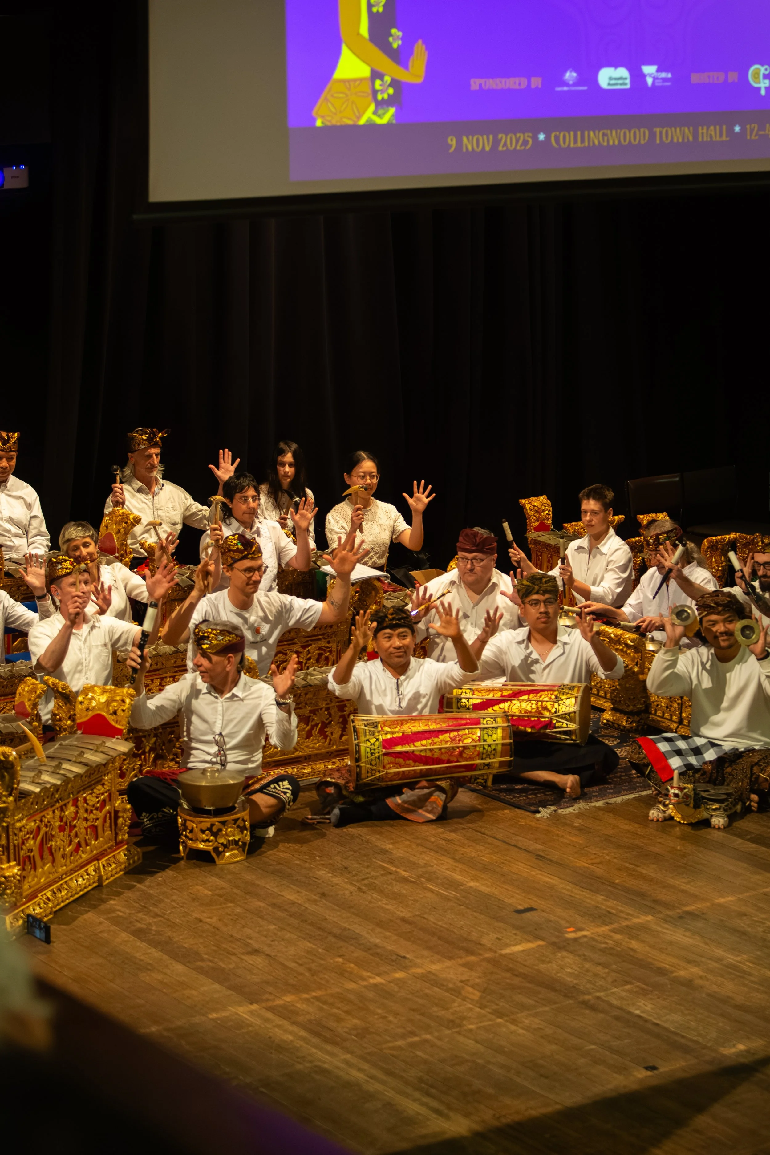 Traditional Indonesian Gamelan ensemble performing on stage with musicians playing various instruments and some raising their hands.