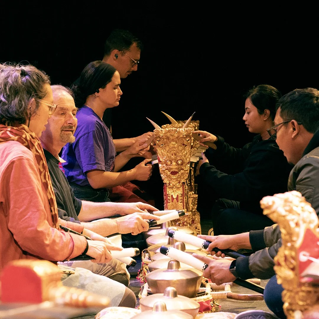 A group of people participating in a traditional cultural ceremony, sitting in a row behind a golden ornamental altar or throne, with musical instruments in front, engaged in a ritual.