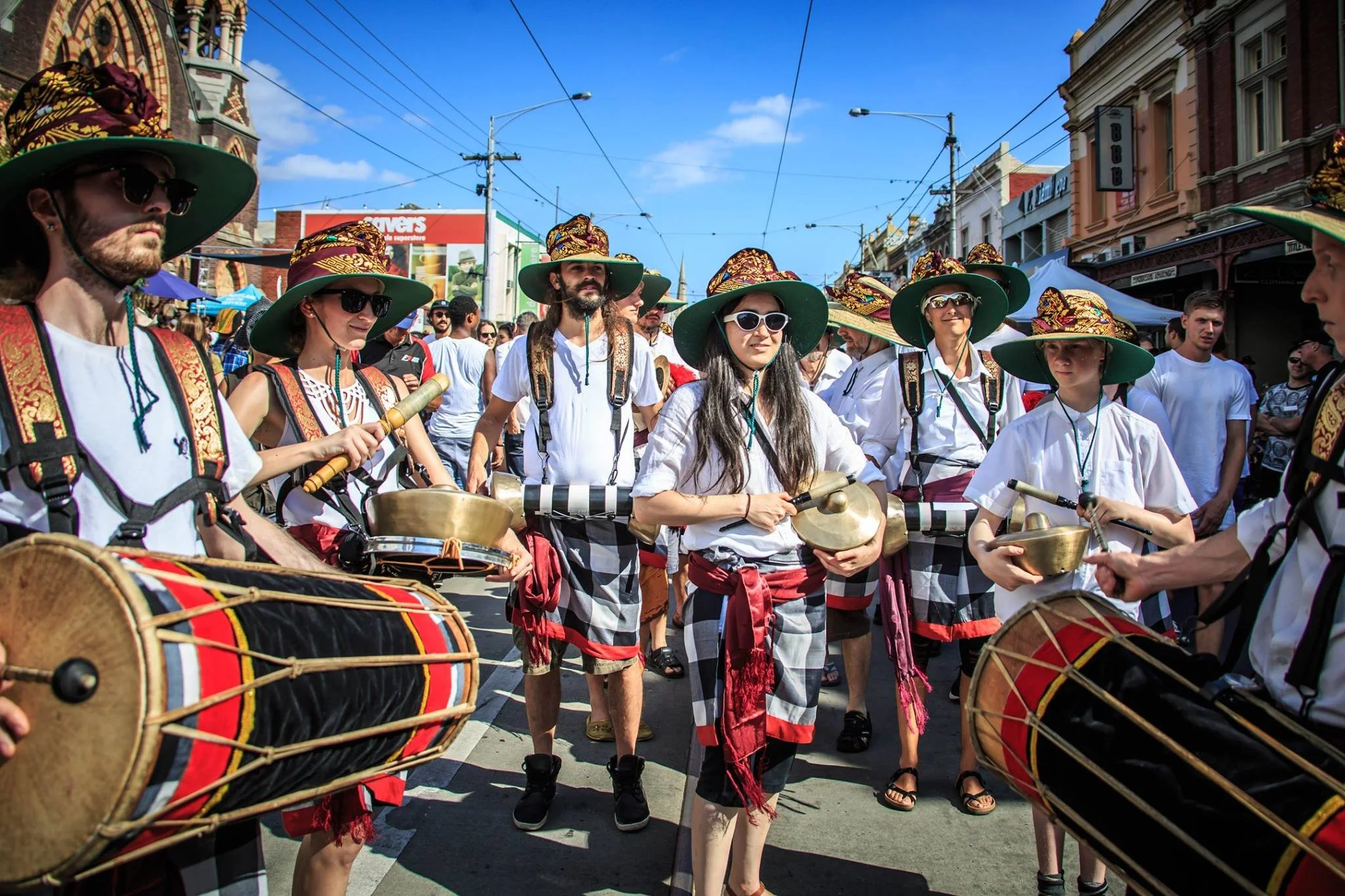 People in a street parade wearing colorful hats, sunglasses, and traditional clothing, playing percussion instruments and drums.
