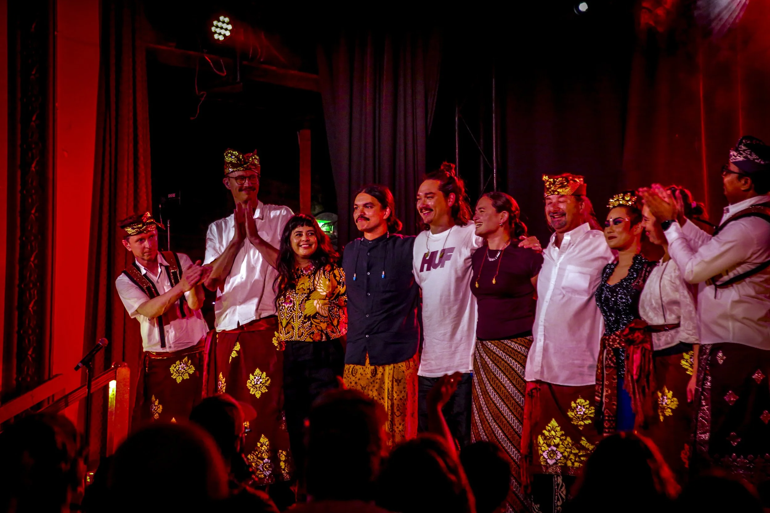 Group of performers on stage after a cultural performance, some wearing traditional Indonesian attire, smiling and engaging with each other, with audience members visible in the foreground.