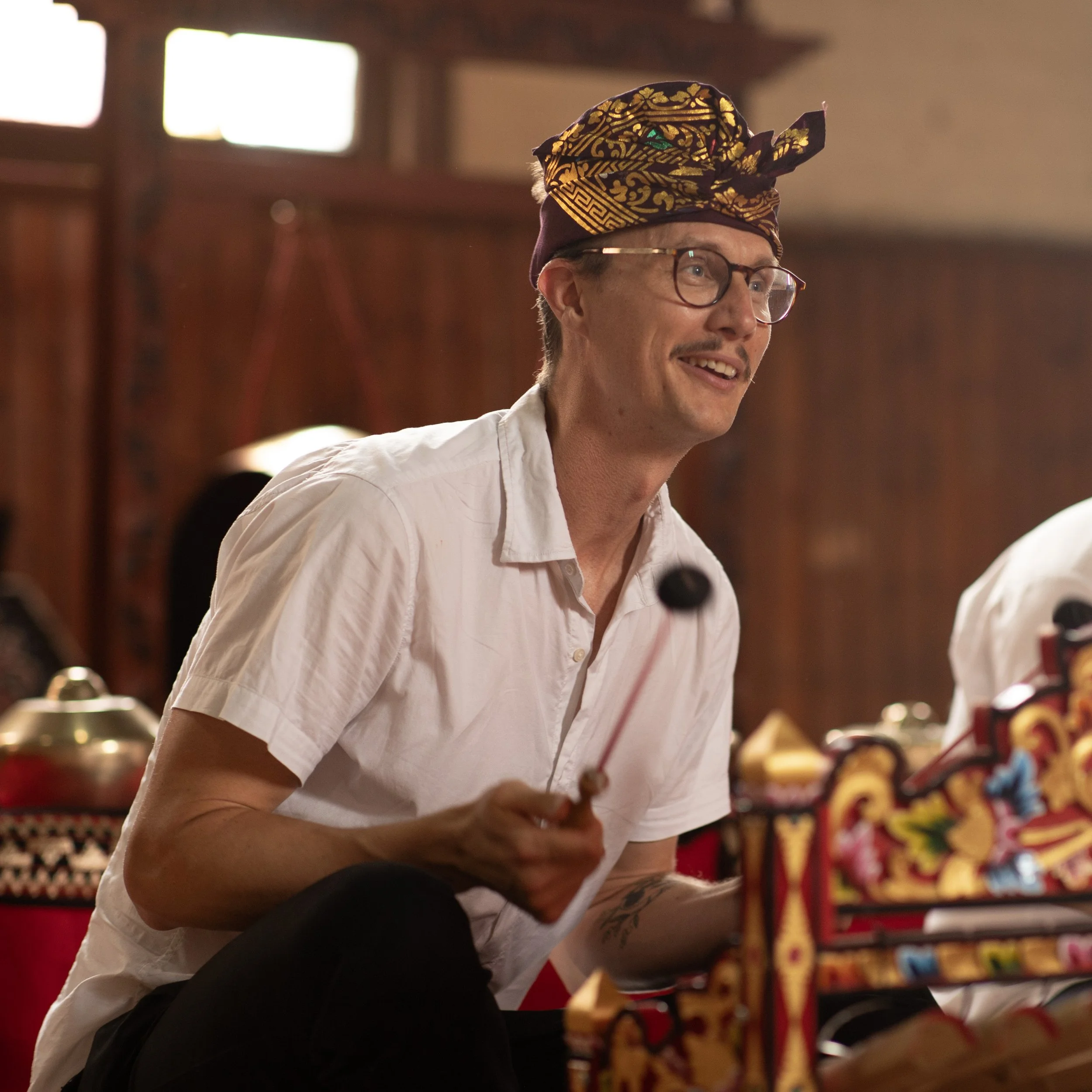 A man with glasses, a white shirt, and a colorful traditional headpiece playing a percussion instrument in a decorated indoor setting.