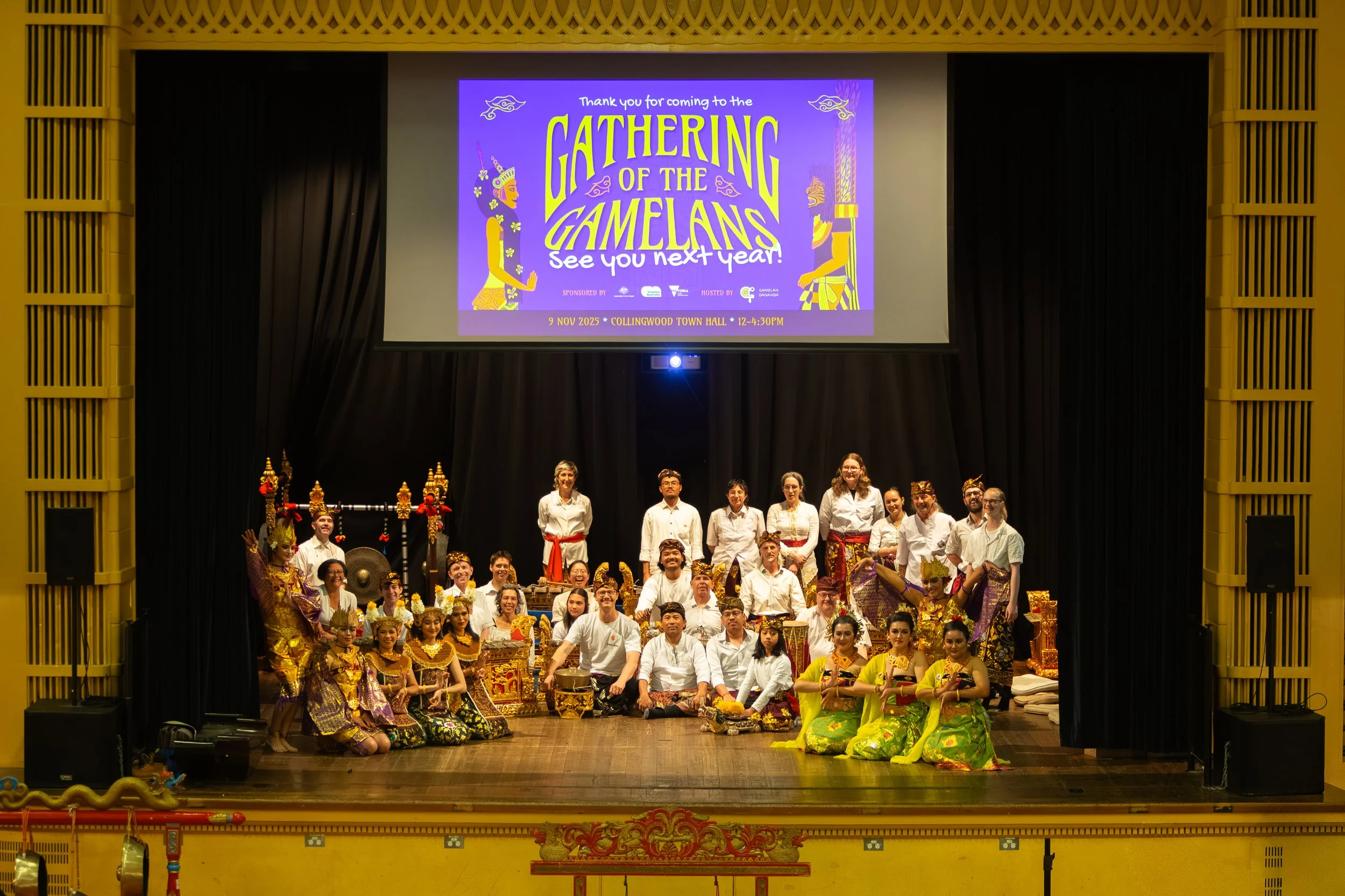 Group of performers in traditional costumes and casual white shirts and pants posing on a stage after a cultural event, with a large digital screen overhead displaying an event announcement for 'Gathering of the Gamelans'.